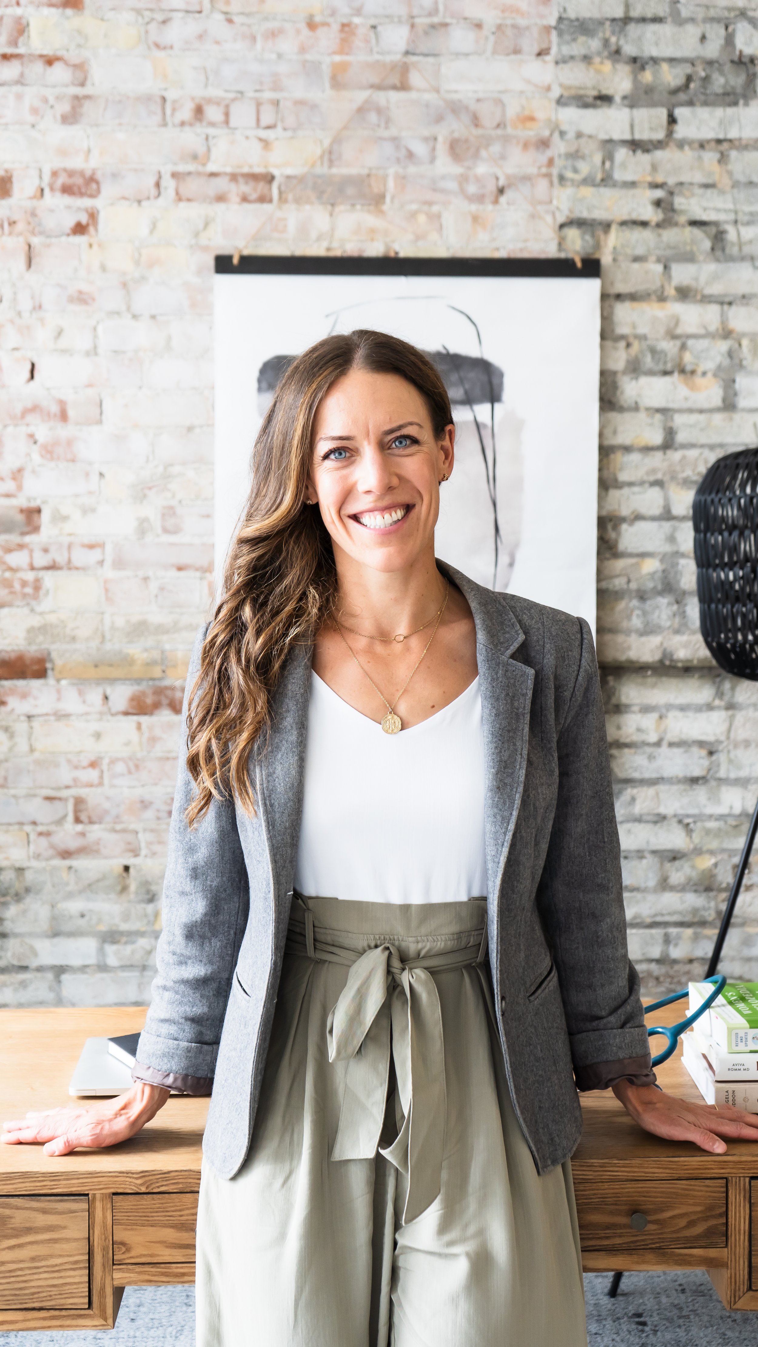 A naturopathic doctor from Kitchener Ontario Canada leaning back on a wooden desk with her hands behind her. She's smiling confidently at the camera. A brick wall is in the background.