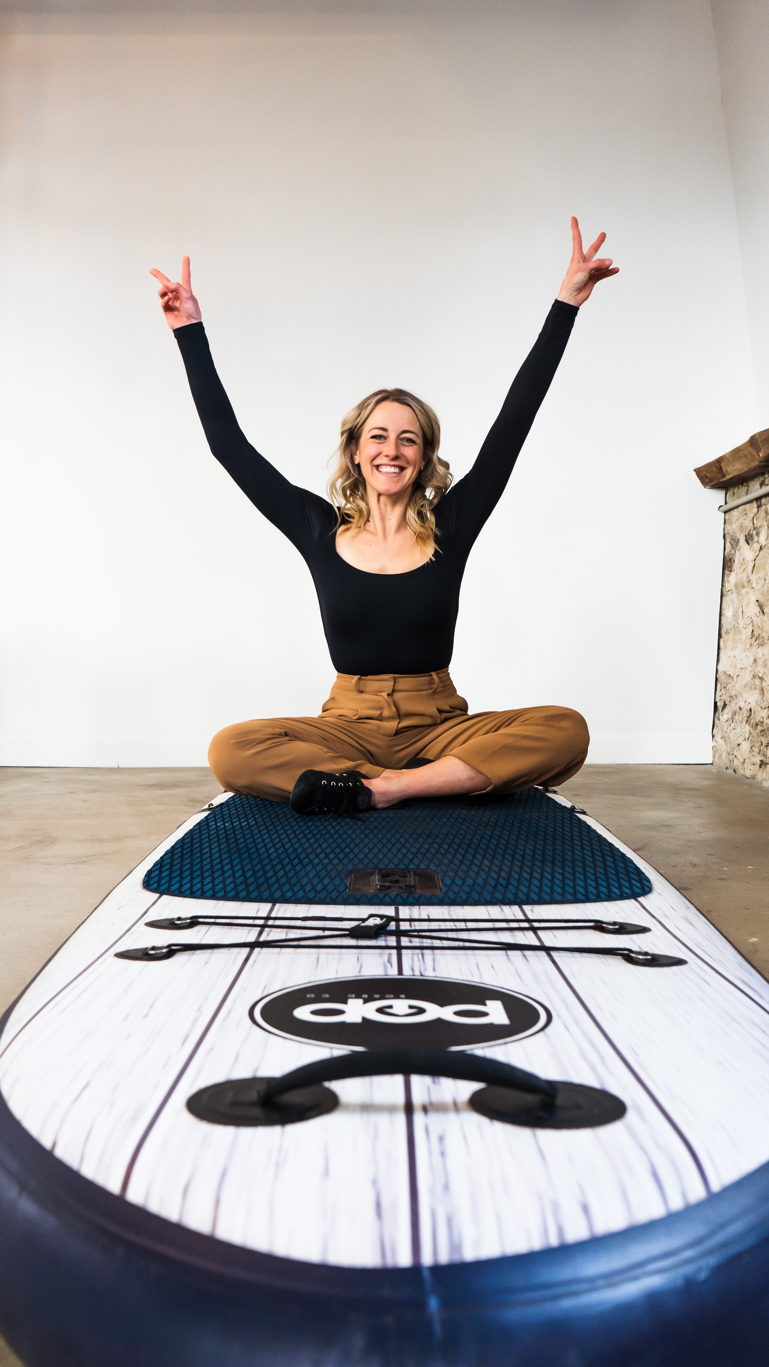 A marketing coach and consultant from Paris Ontario Canada posing on her paddle board, sitting cross legged with her hands up in the air, smiling confidently at the camera.