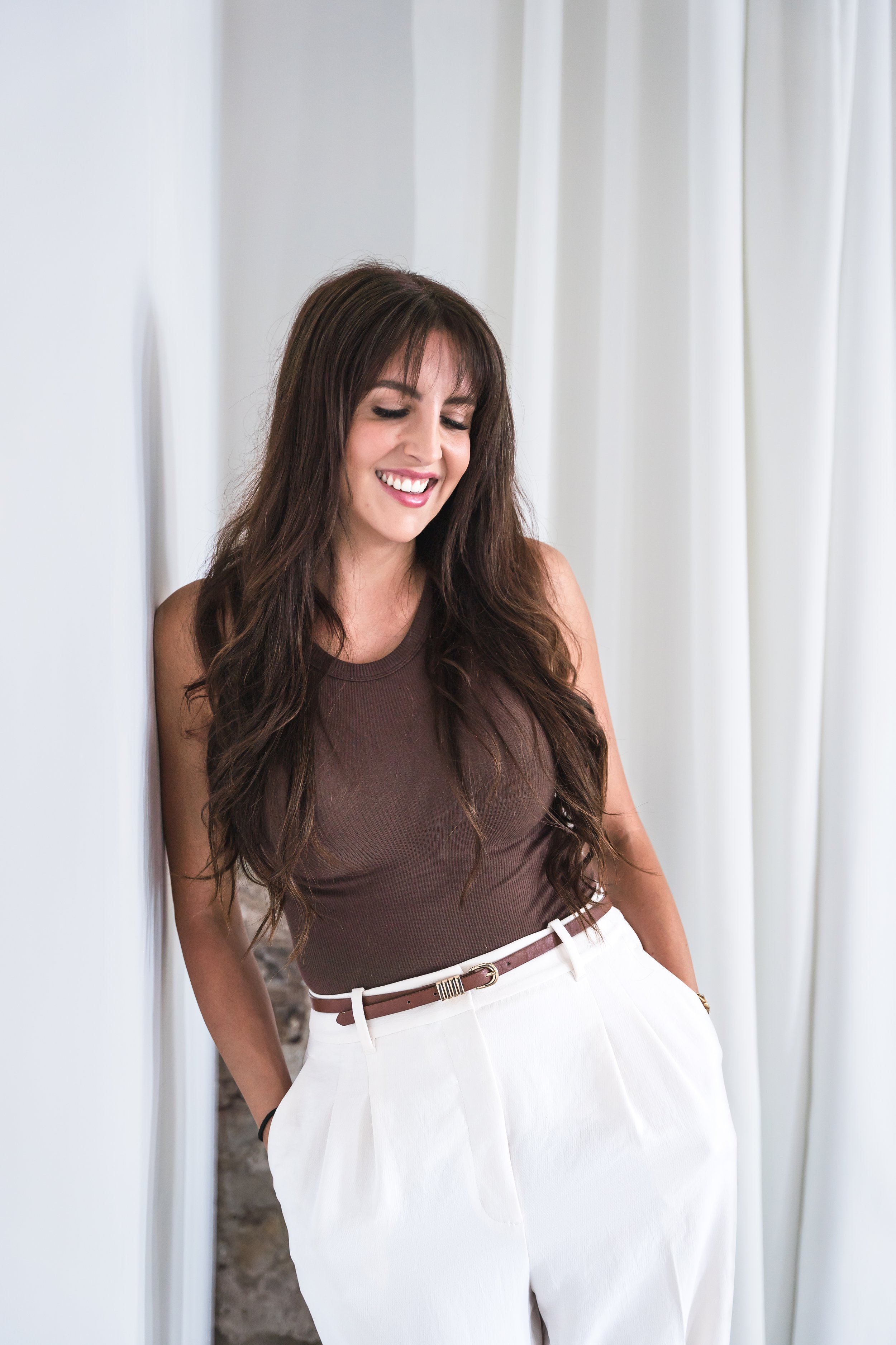 A medical aesthetician from Brantford Ontario Canada posing leaned up against a white wall with her hands in the pockets of her trousers. She's smiling down at the floor.