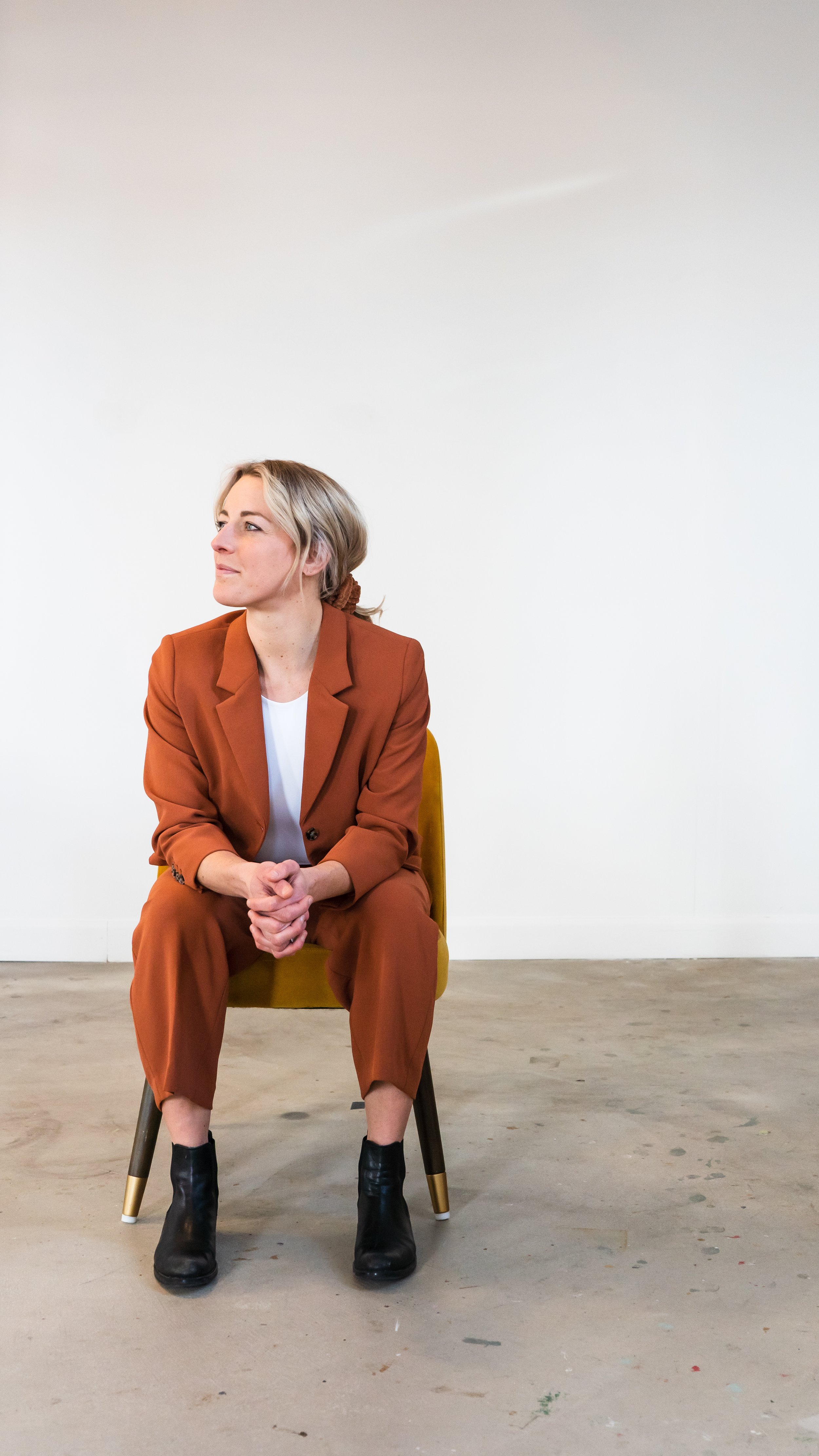 A marketing coach and consultant from Paris Ontario Canada posing in front of a white wall. She's sitting on a chair leaning on her knees, looking off to the side.