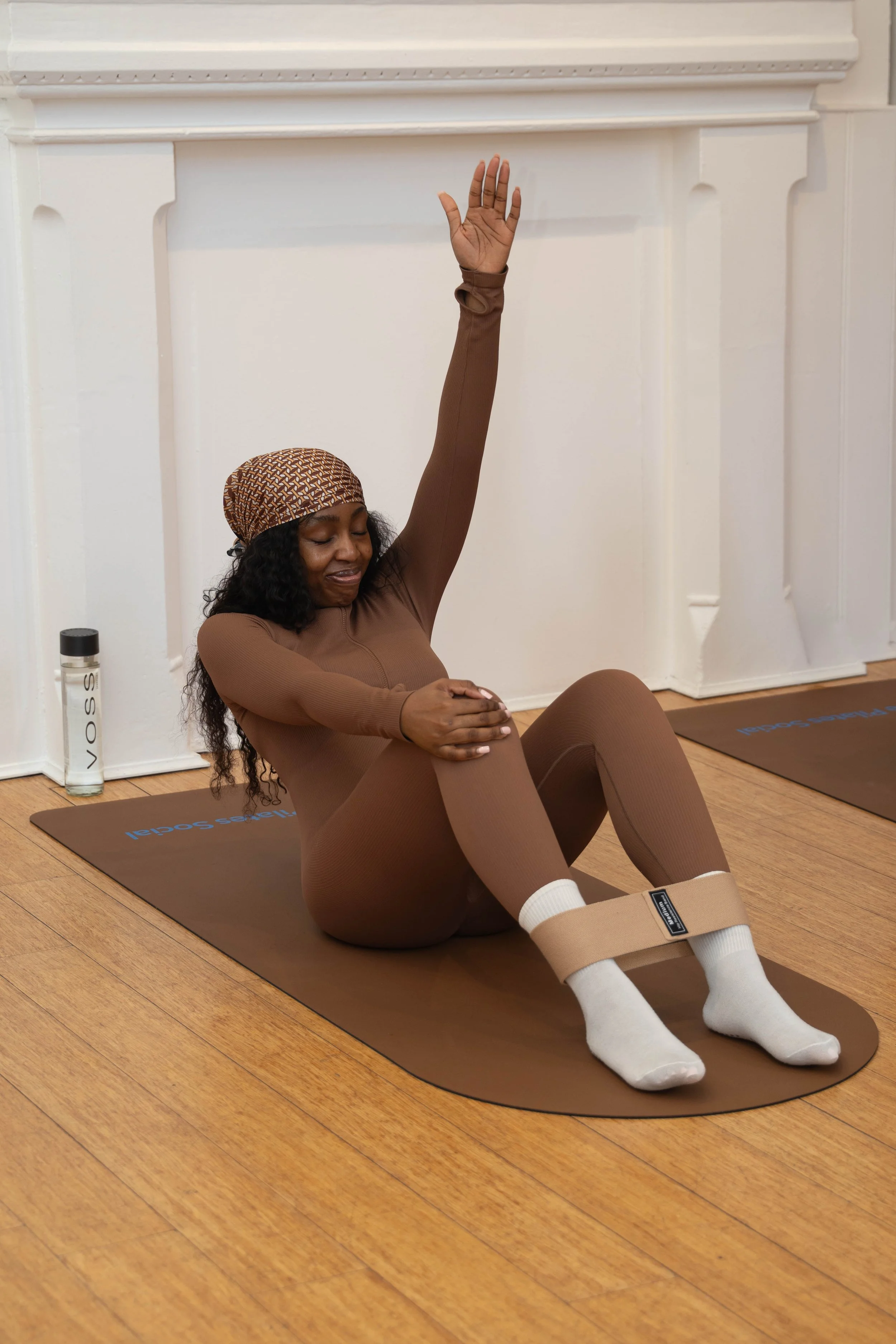 Woman in wellness attire sitting on a yoga mat, raising one arm and holding her knee in a seated exercise, with a serene facial expression and face covered with a headscarf.