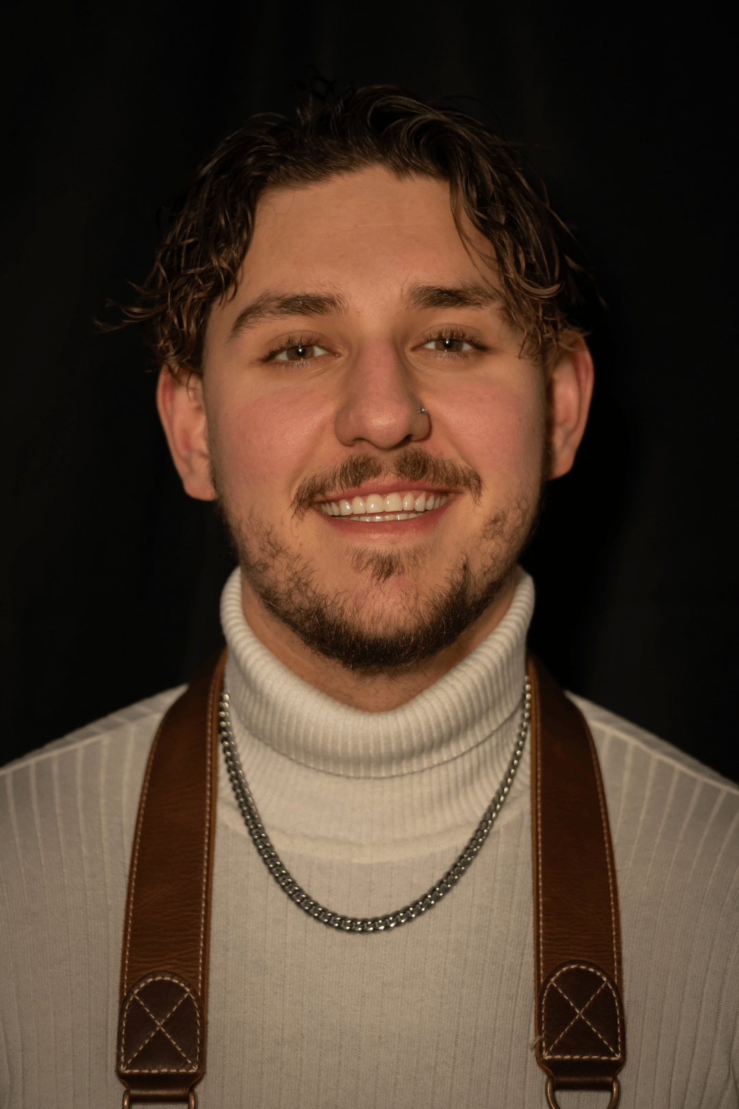 A young man with curly brown hair, a nose piercing, and facial hair wearing a white turtleneck sweater, a silver chain, and leather suspenders, smiling against a black background.