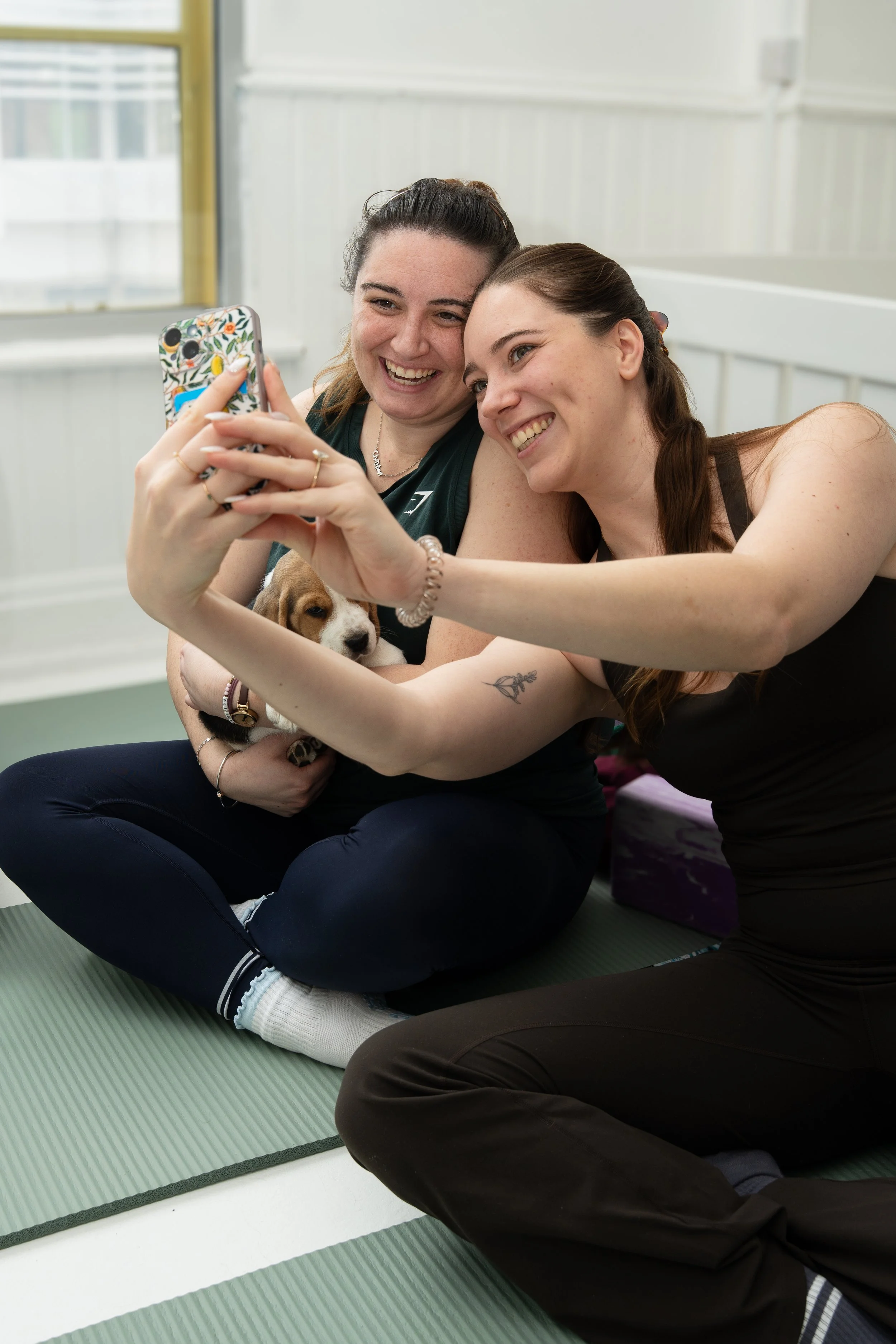 Two women taking a selfie with a puppy, sitting on a green exercise mat in a well-lit room with white walls and large windows.