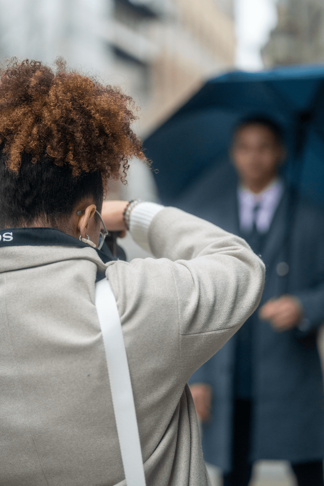 A person with curly hair taking a photo of another person holding an umbrella in an urban setting.