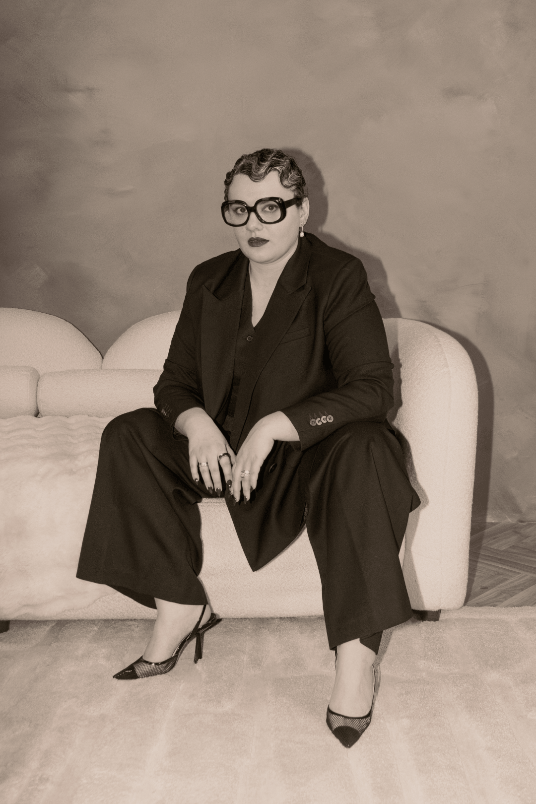 A woman sitting on a sofa, wearing a black suit, large glasses, and high heels, with a plain background in a vintage style.