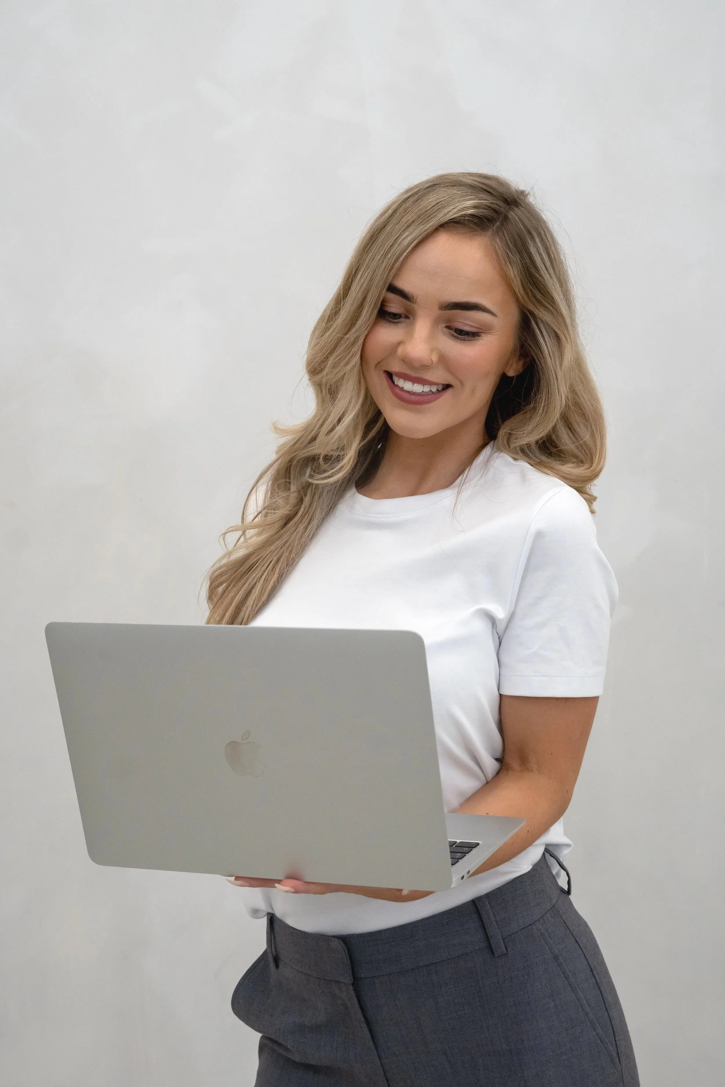A woman with blonde hair smiling and looking at a silver laptop, dressed in a white t-shirt and gray pants, standing against a plain white wall.