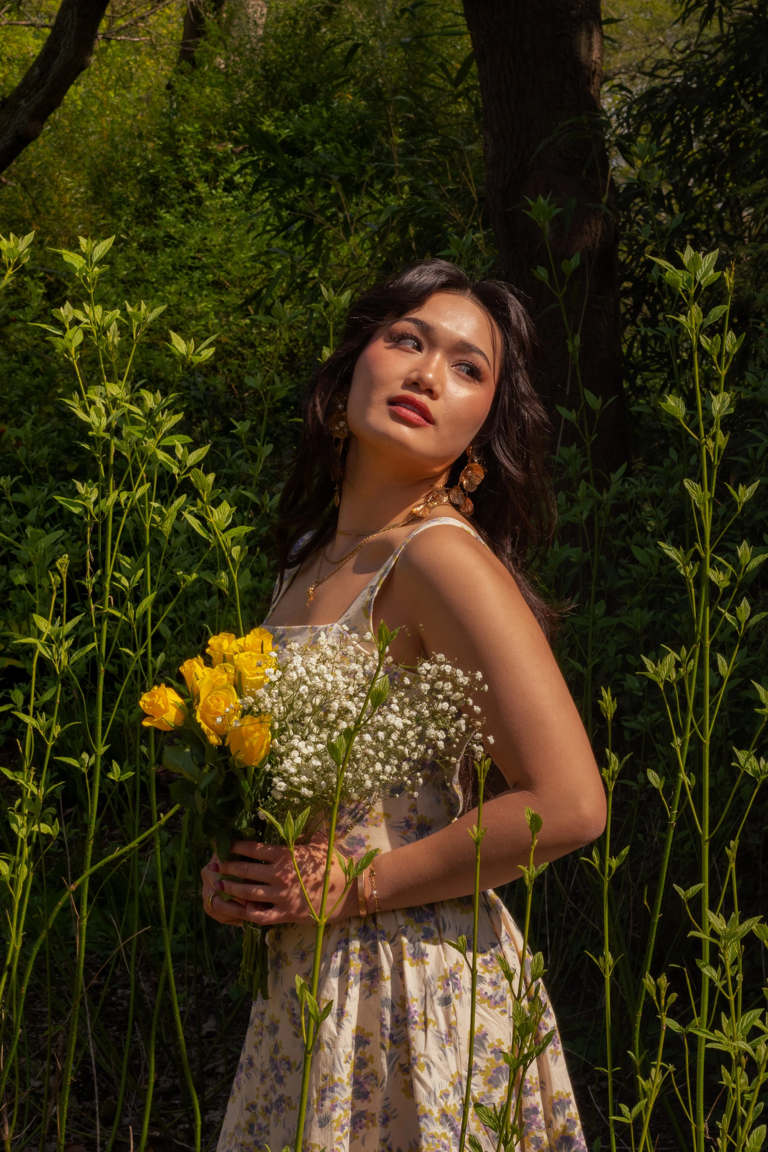 A woman with dark hair and makeup holding a bouquet of yellow and white flowers, standing in a lush green forest with tall plants and trees surrounding her, sunlight filtering through the foliage.