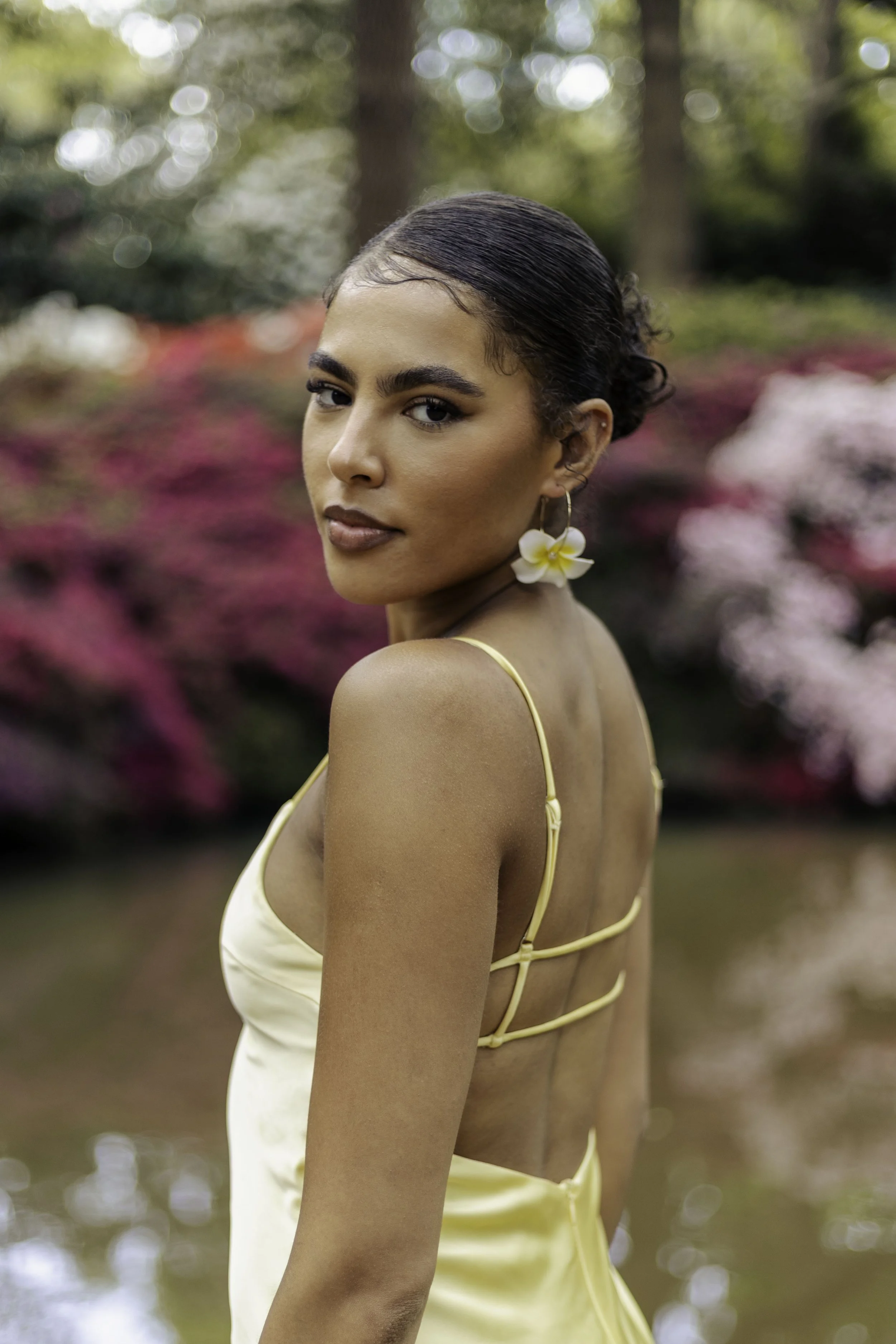 A woman with dark, slicked-back hair, wearing a yellow, strappy dress and a white flower earring, posing outdoors with a blurred background of trees and pink flowers.