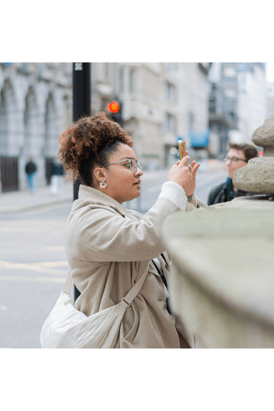 A woman with curly hair in a beige coat and glasses takes a photo with her smartphone on a city street, with buildings and a red traffic light in the background.