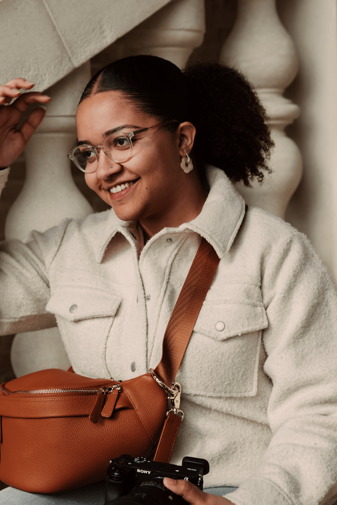 Woman with glasses, earrings, and a nose piercing, smiling, holding a camera with a brown crossbody bag, indoors near a stone railing.