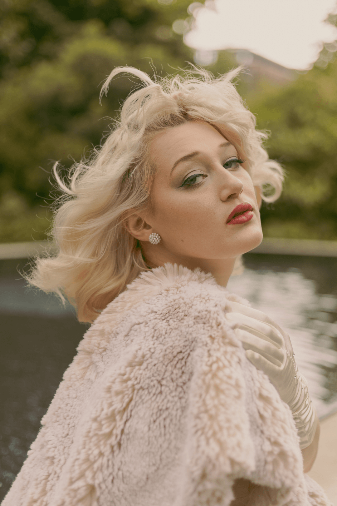 A woman with platinum blonde hair styled in soft waves, wearing pearl earrings, red lipstick, a white faux fur coat, and gloves, standing outdoors near a body of water with blurred greenery in the background.