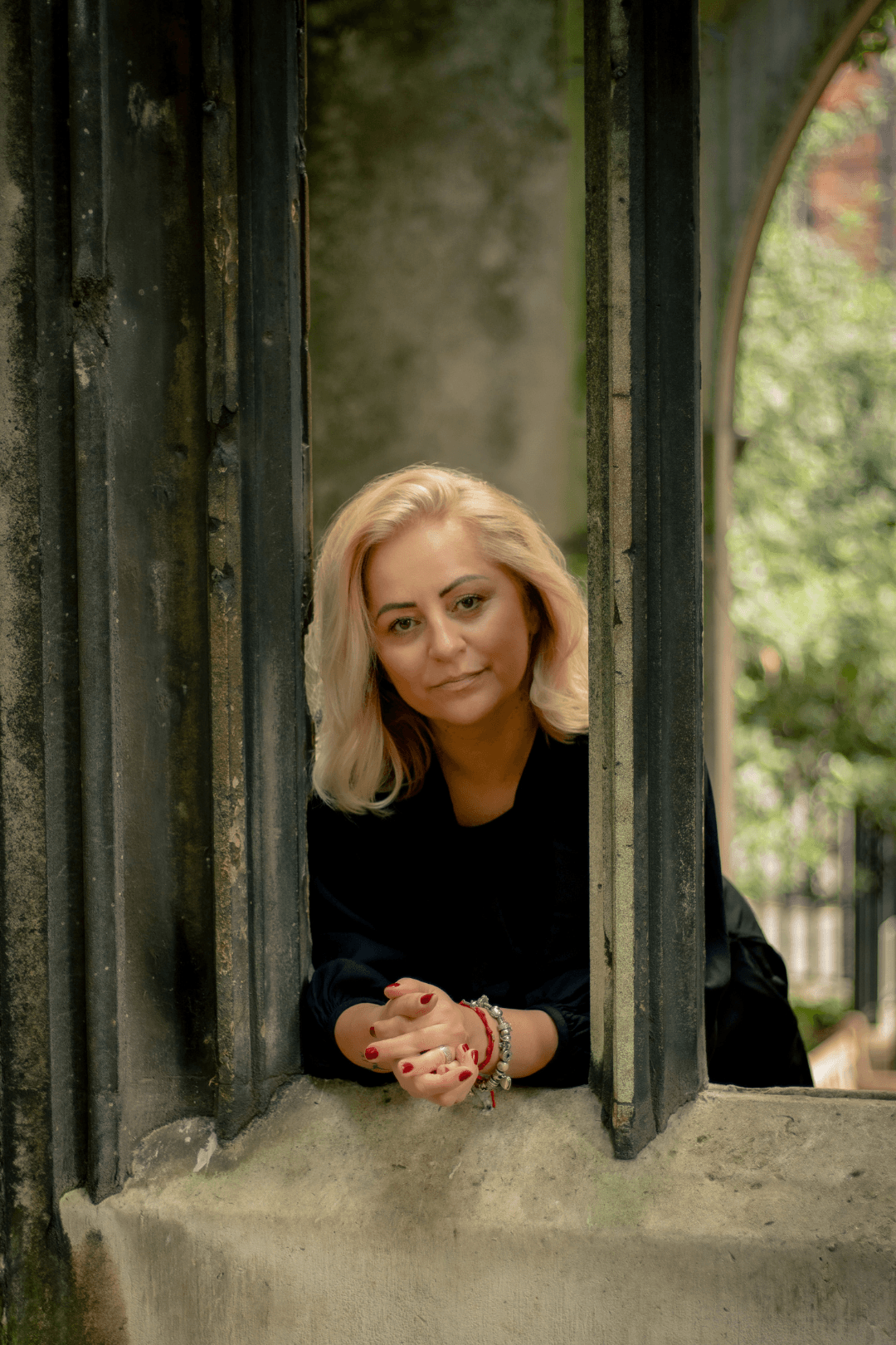 Woman with blonde hair leaning out of a window with a grey stone frame, greenery in the background.
