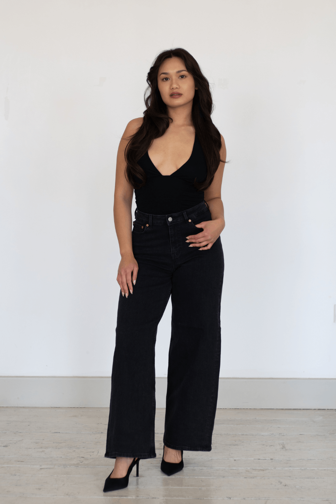 A woman with long, wavy dark hair wears a black sleeveless V-neck top and high-waisted black jeans with black high heels, standing against a plain white wall.