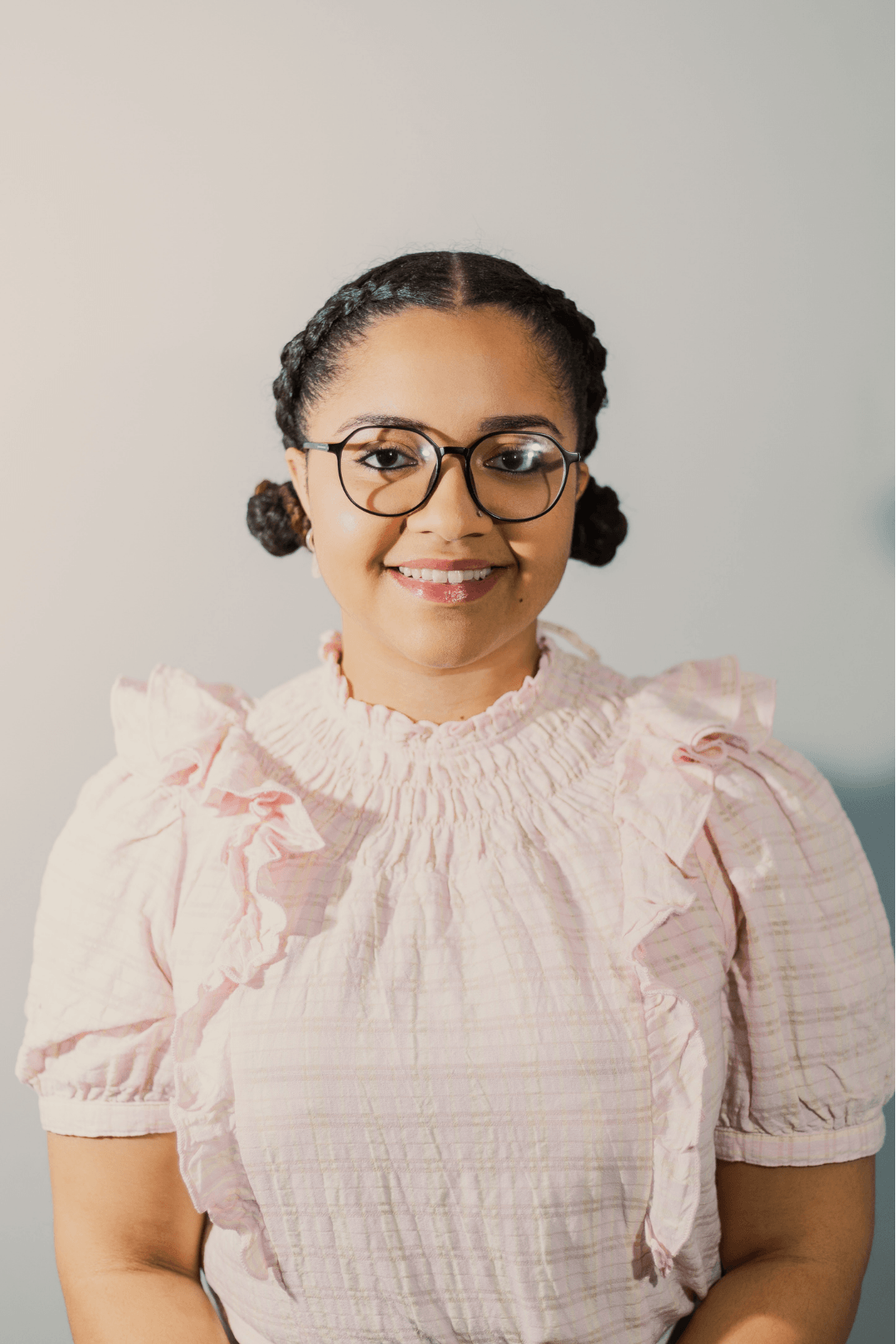 A young woman with glasses, dark hair styled in braided buns, wearing a pale pink, ruffled blouse, and smiling.