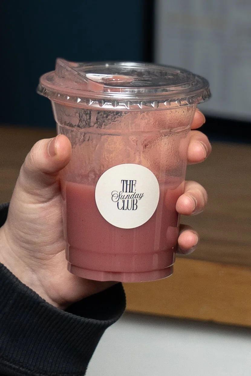 A hand holding a clear plastic cup with pink-colored drink and a sealed lid, labeled "The Sunday Club".