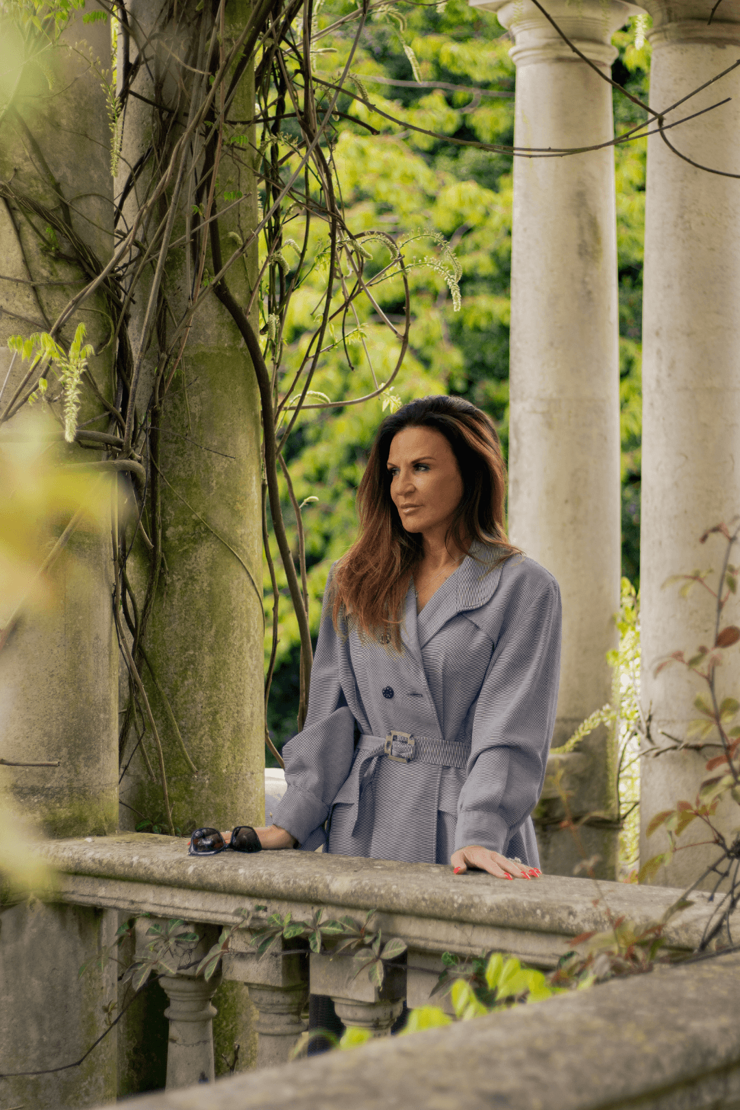 A woman in a gray trench coat leaning on a stone railing outdoors, with ancient columns and lush greenery in the background.