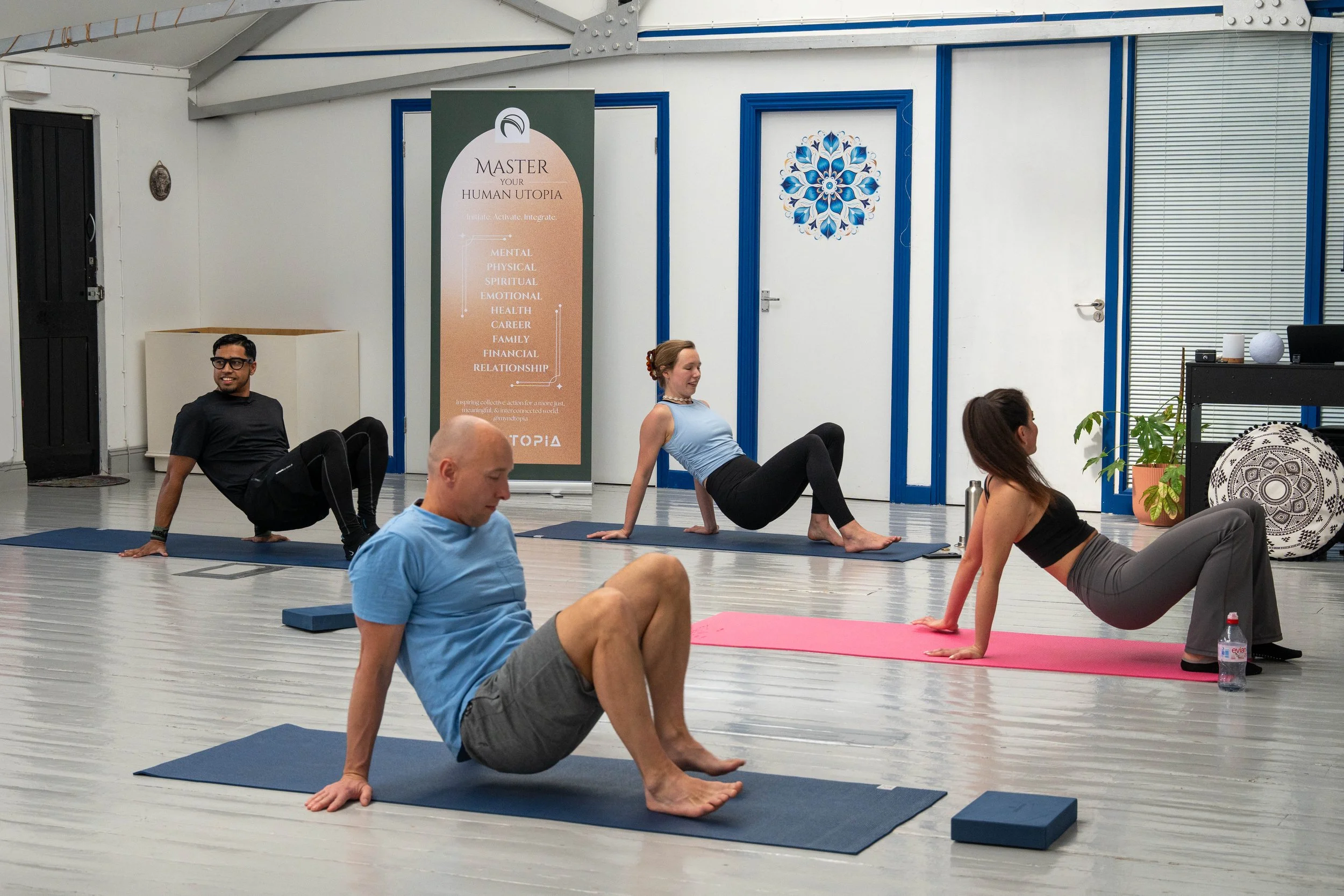 People participating in a yoga or fitness class in a bright room with a sign that reads "Master Your Human Utopia" and lists areas like mental, physical, spiritual, emotional, health, career, family, financial, and relationship. Participants are on m