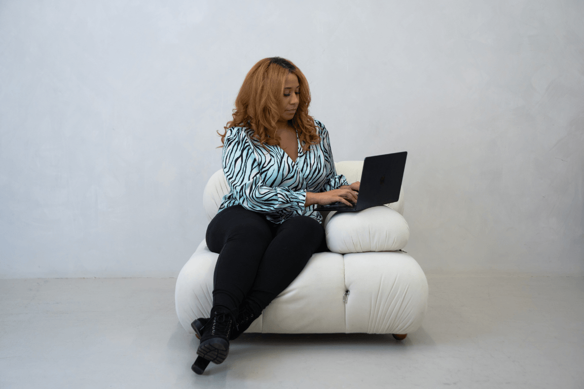 A woman with curly, reddish-brown hair sitting on a white couch, working on a black laptop, against a plain light gray wall.