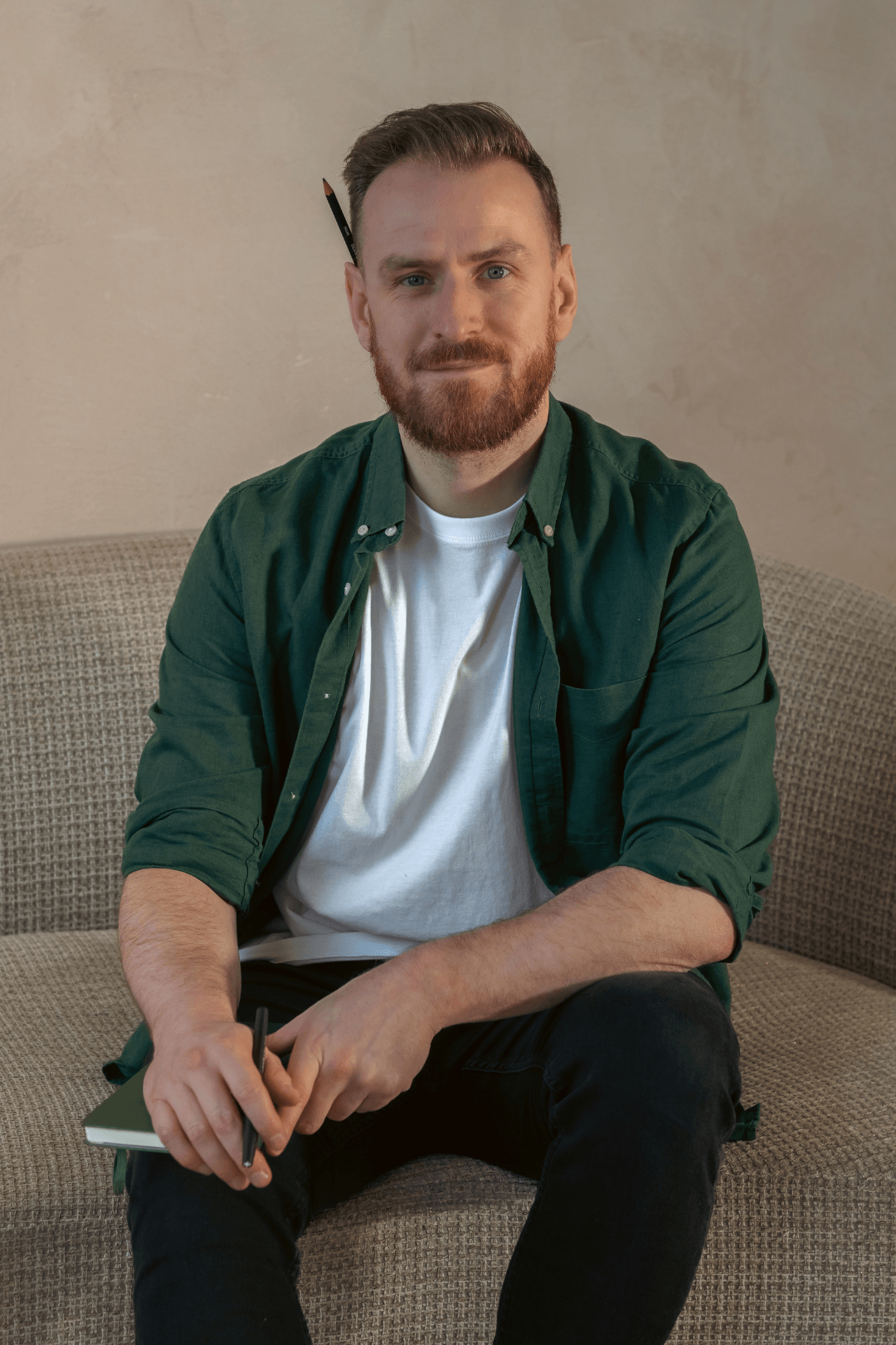 Young man with a beard and light brown hair, sitting on a beige textured sofa, with a pencil behind his right ear, wearing a white t-shirt and a green button-up shirt.