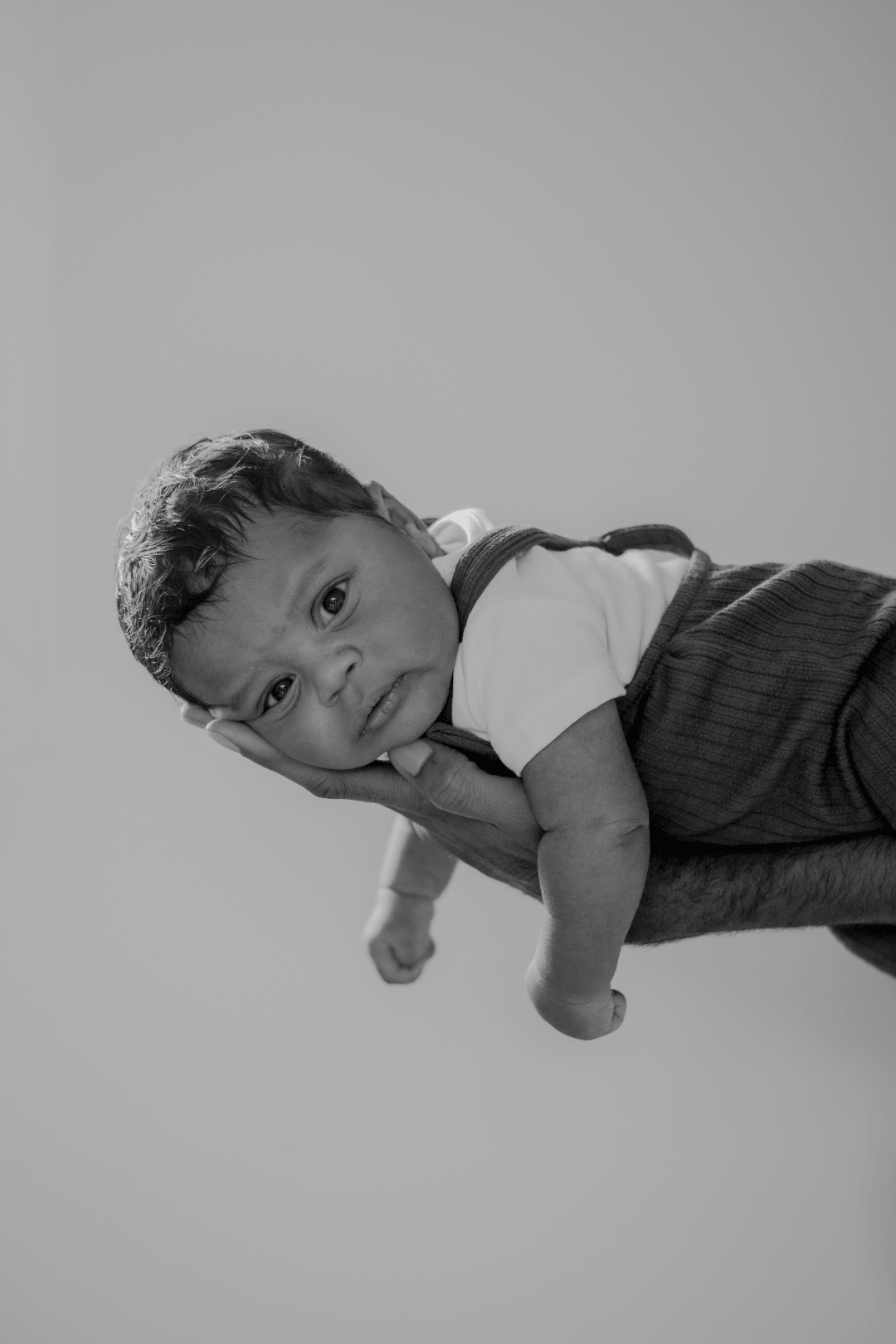 A person holding a baby boy in the sky, with the baby resting on their palm. The baby looks curious and is wearing a white shirt and dark overalls, and the image is in black and white.