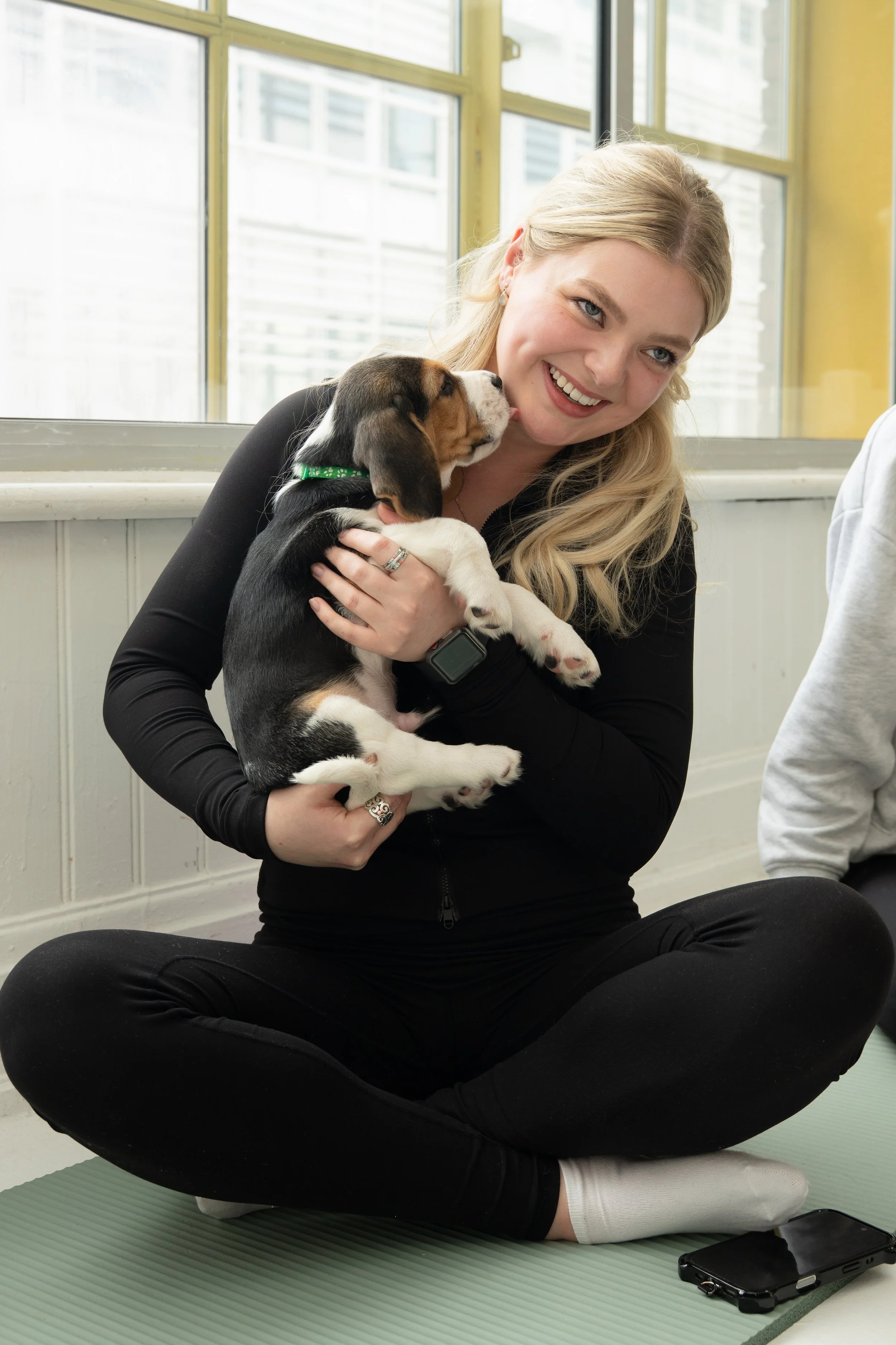 A young woman with long blonde hair smiles while sitting cross-legged on a yoga mat, holding a small beagle puppy. The puppy is licking her face. She is wearing a black zip-up jacket and white socks. There is a phone on the mat beside her, and the ba