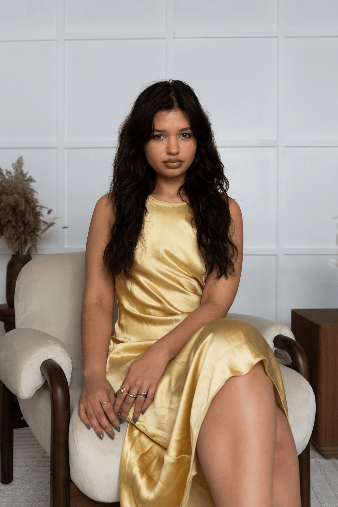 A young woman with long wavy dark hair and tan skin sits on a cream-colored armchair, wearing a shiny yellow satin dress, in a minimalist room with white grid-patterned walls and a wooden side table with a dried plant.