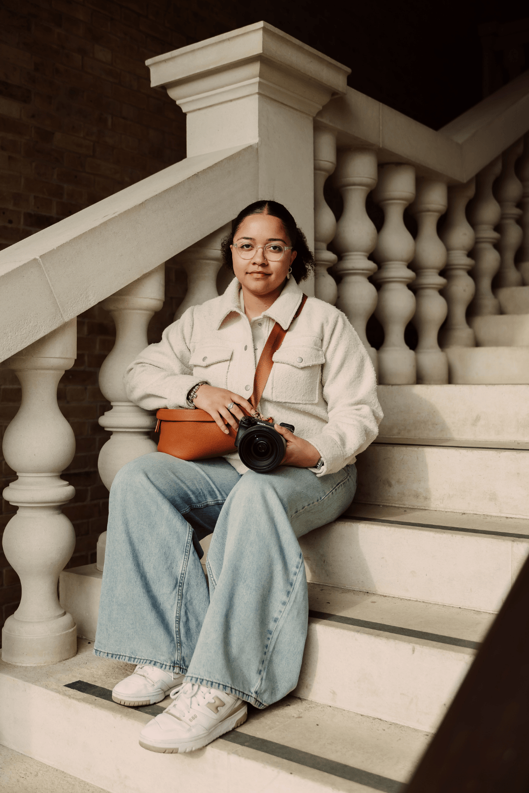 A woman sitting on stairs holding a camera, wearing glasses, a white jacket, light blue jeans, and white sneakers, with a brown bag and a watch, in front of a brick wall and ornate columned railing.