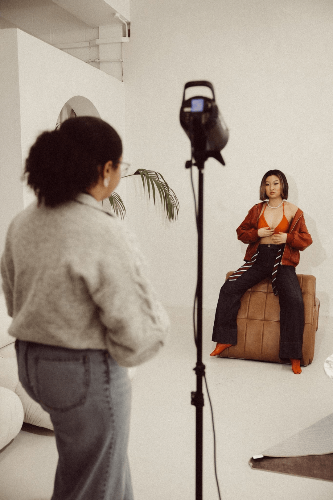 A woman with shoulder-length hair sitting on a brown ottoman, wearing an orange top, black pants, a red jacket, orange high heels, and jewelry during a photoshoot in a studio.