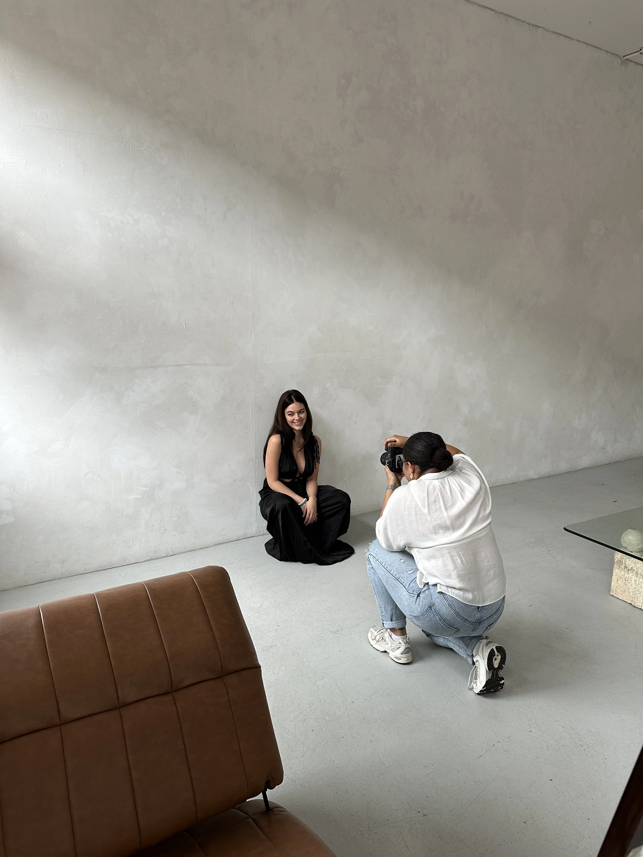 A woman in a black dress sits on the floor as a photographer kneels in front of her, taking her picture against a plain white wall.