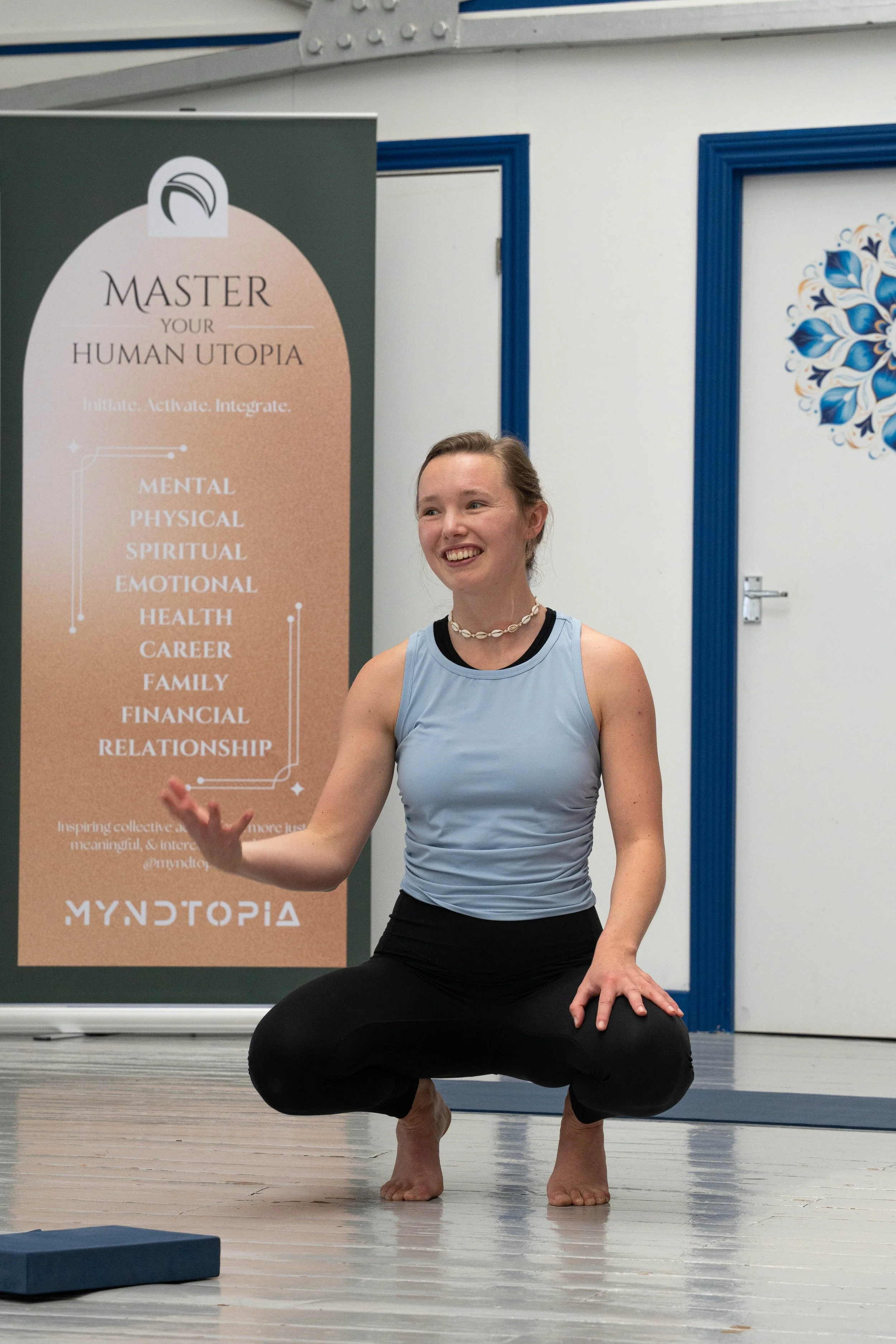 A woman practicing yoga indoors, squatting on a wooden floor with her hands resting on her thighs, wearing a light blue athletic top and black pants, smiling while looking to her right. A vertical poster with the words "Master Your Human Utopia" and 