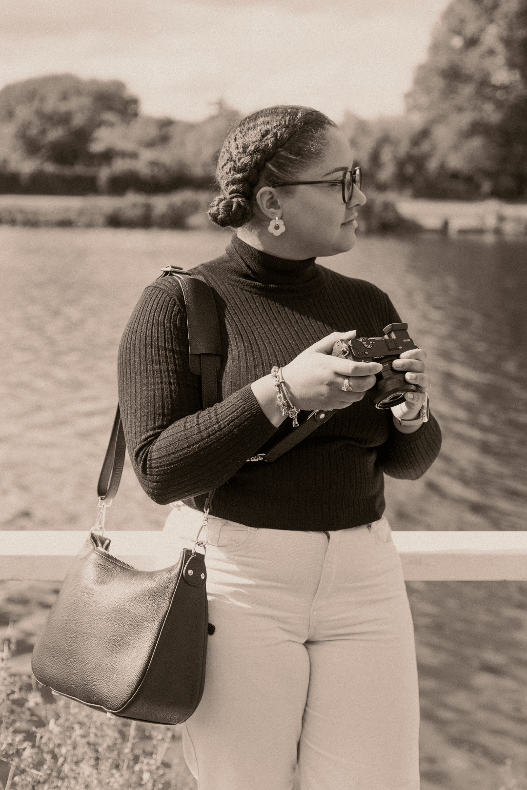 A woman with braided hair, glasses, and earrings holding a camera outdoors by a body of water, wearing a black turtleneck and light-colored pants.