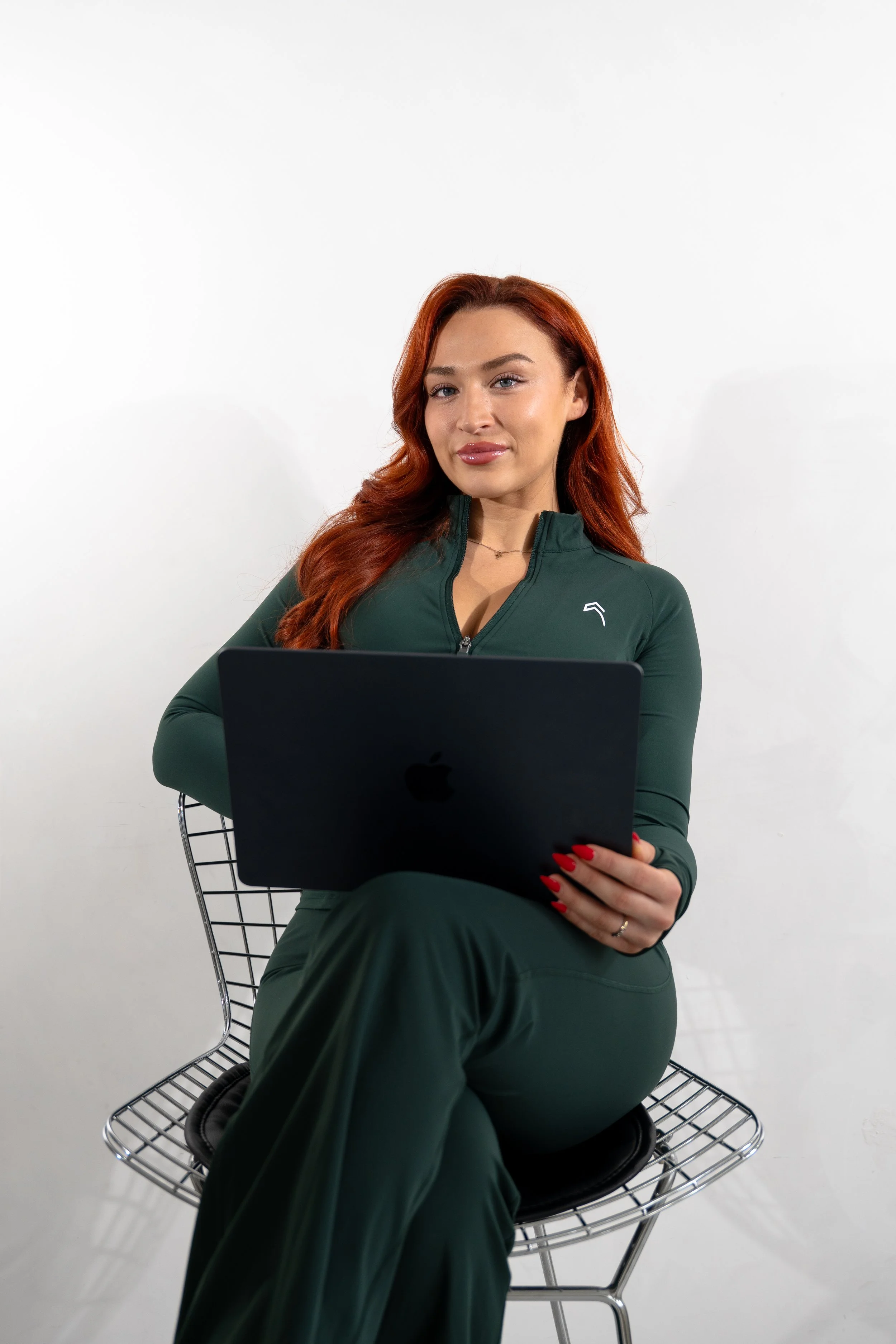 A young woman with long red hair, wearing a green gym set, sitting on a metal chair with a white background, holding her laptop and smiling at the camera.