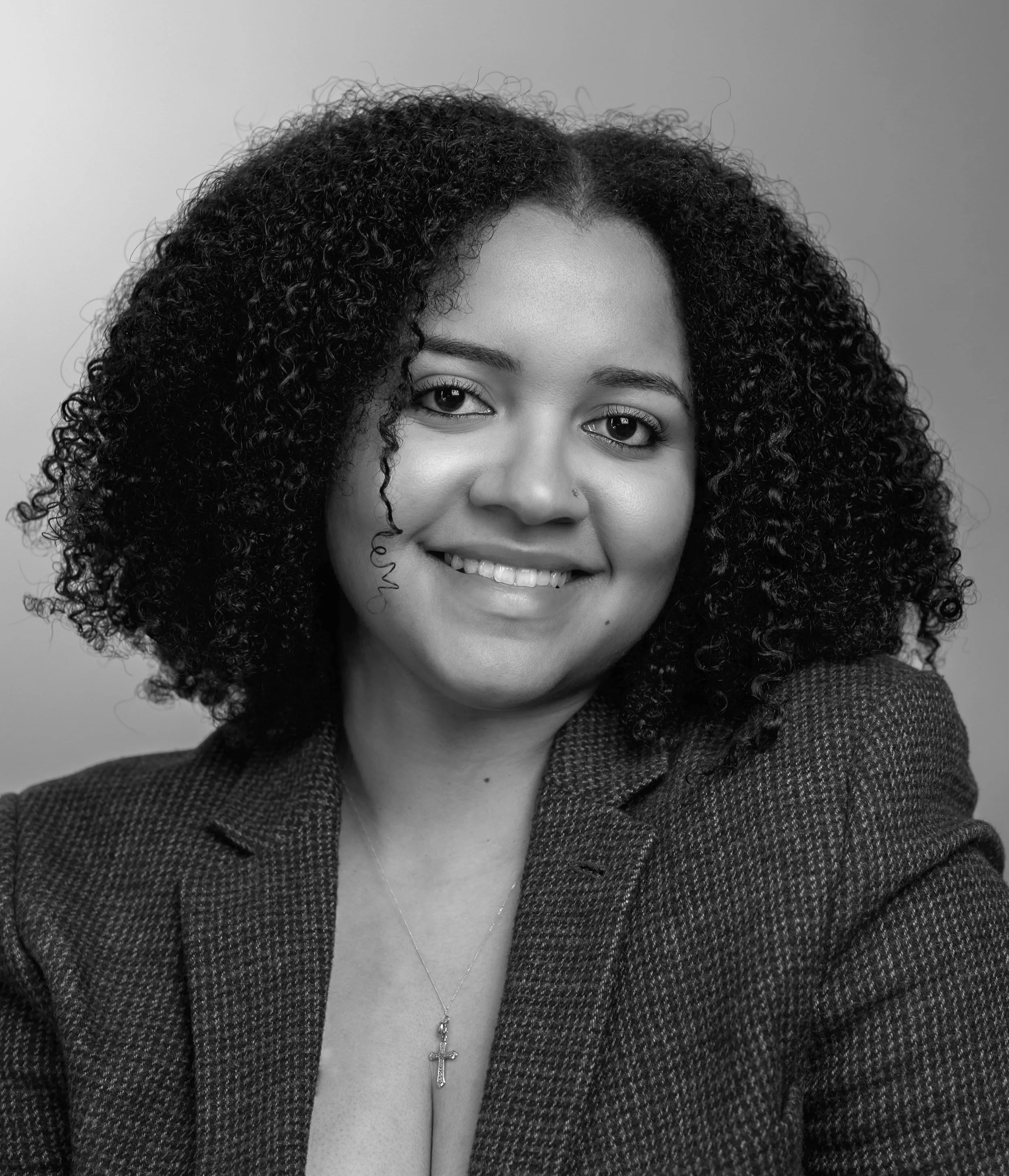 A woman with curly hair smiling, wearing a blazer, a necklace with a cross pendant, and a top with a deep neckline.