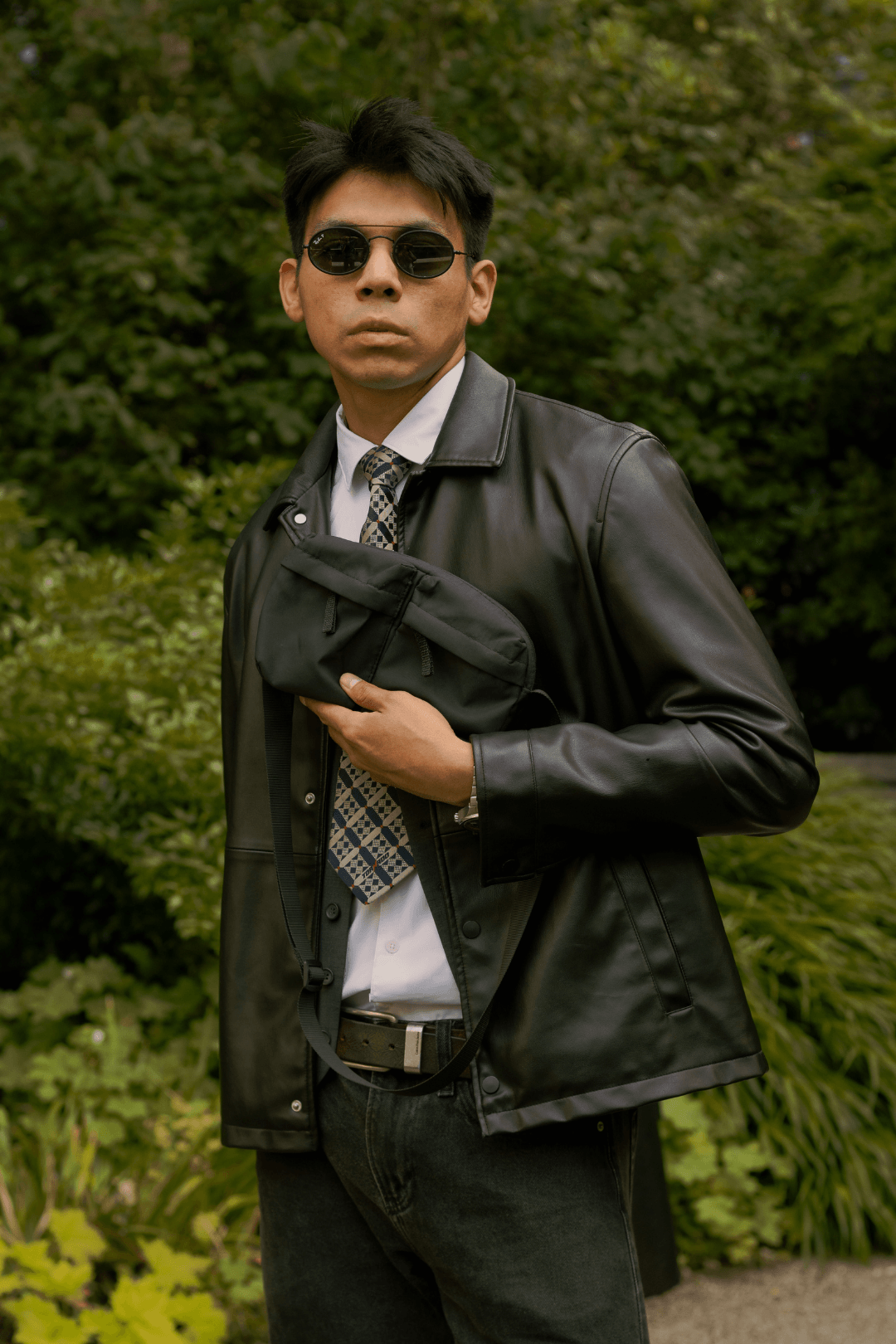 A man wearing sunglasses, a leather jacket, and a tie holding a black bag across his chest outdoors with green foliage in the background.