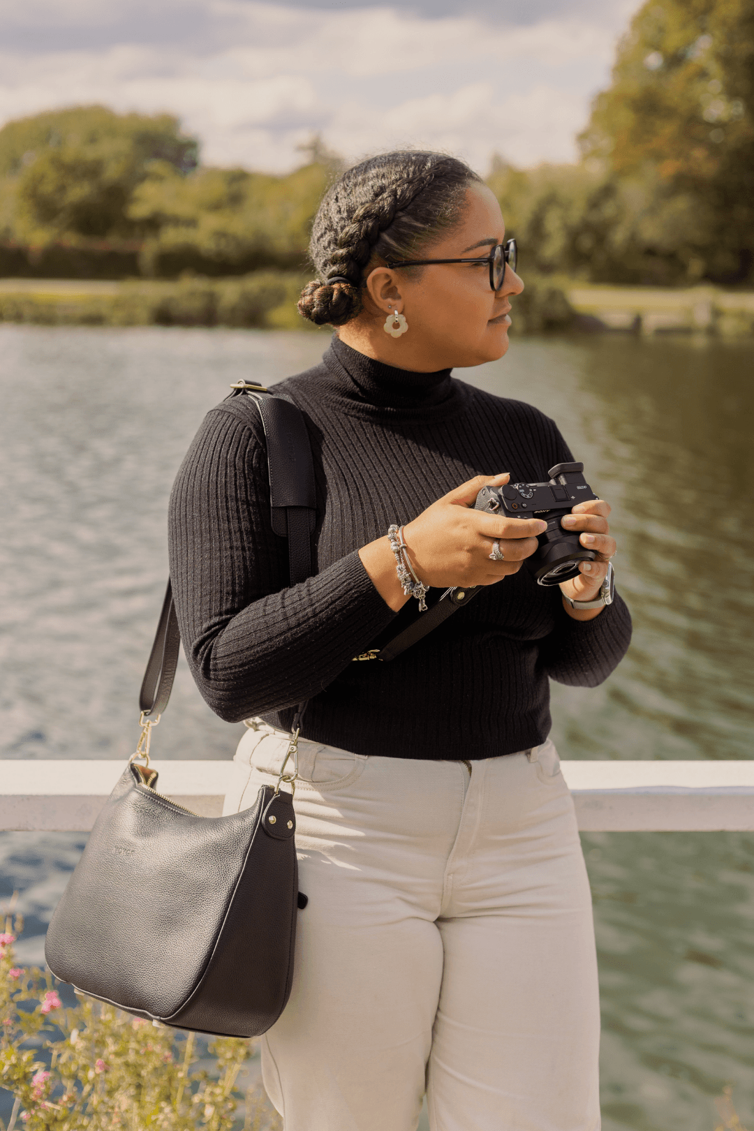 A woman standing by the water holding a camera, wearing glasses, a black turtleneck, and cream-colored pants with a black shoulder bag.