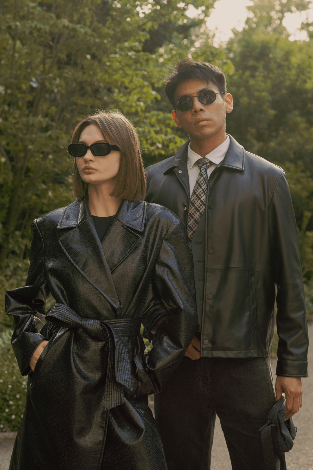 A woman and a man are standing outdoors with trees in the background. They are both wearing black leather jackets and sunglasses, with a serious expression.