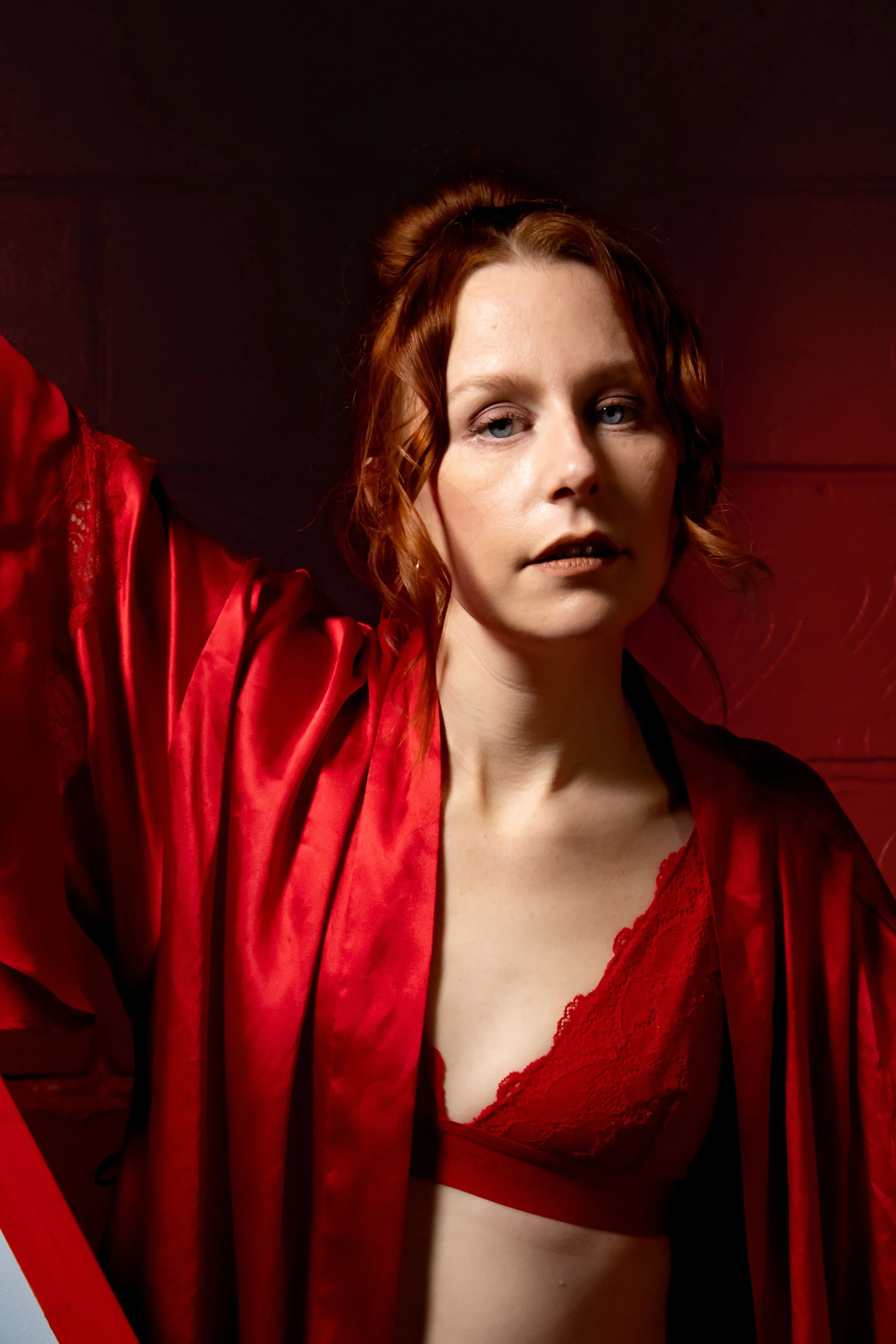 Close-up of a woman with blue eyes and red hair wearing red lingerie and a red silk robe, against a dark background.