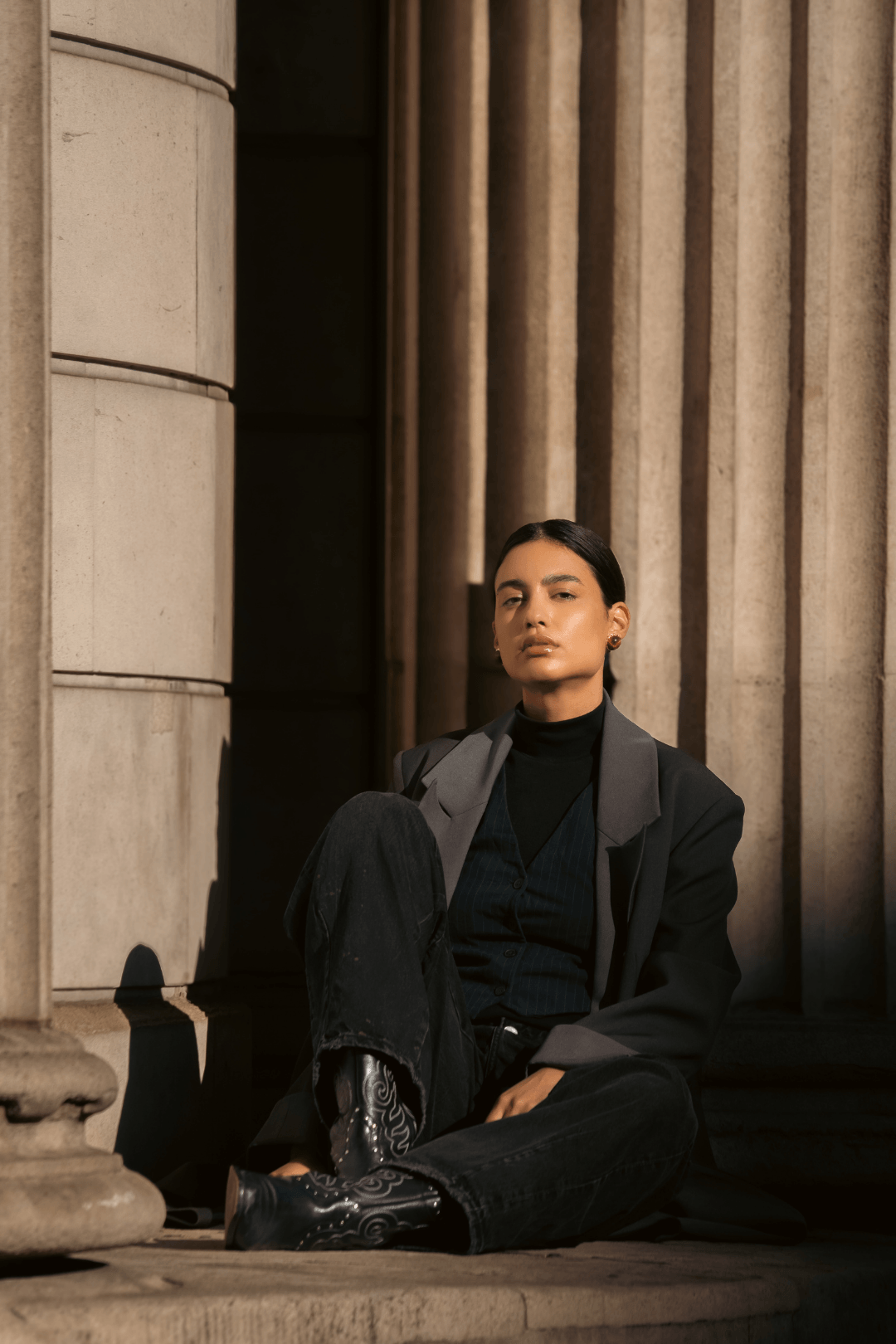 A young woman sitting on the steps of a building with classical architecture, wearing a black top, dark pants, a gray jacket, and cowboy boots, looking confidently at the camera.
