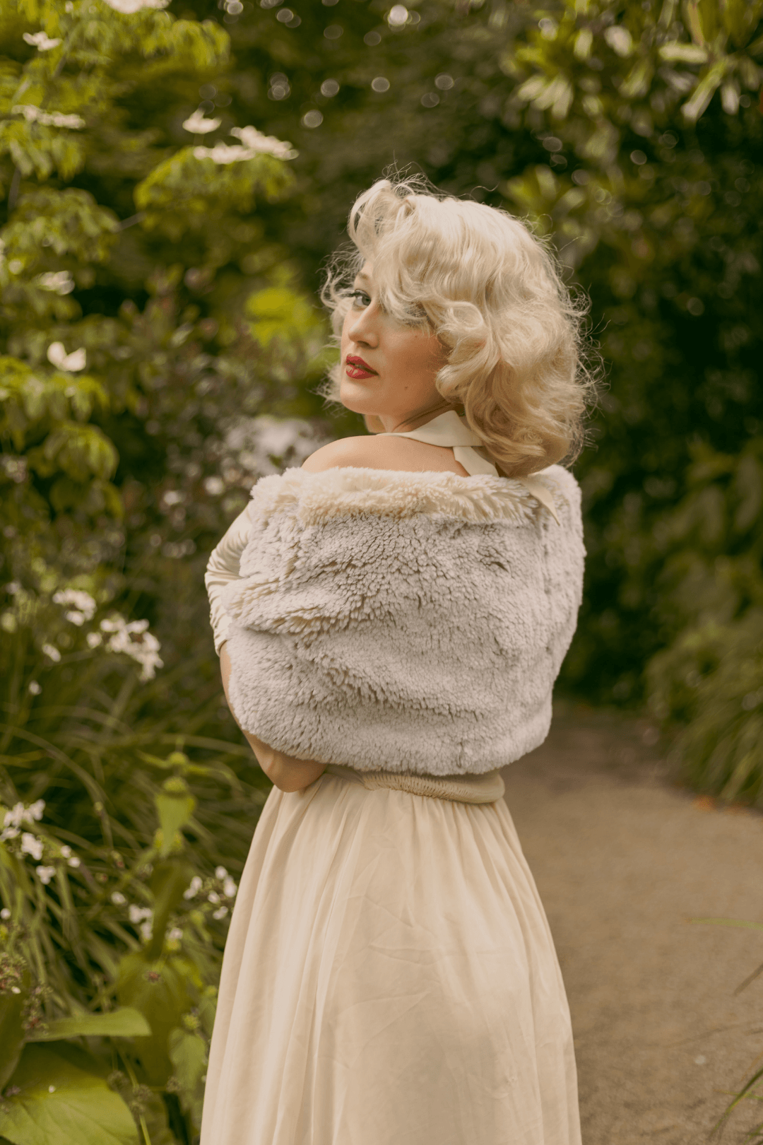 A woman with blonde, curly hair wearing a vintage cream-colored dress and a fur stole standing outdoors on a nature trail surrounded by green foliage.