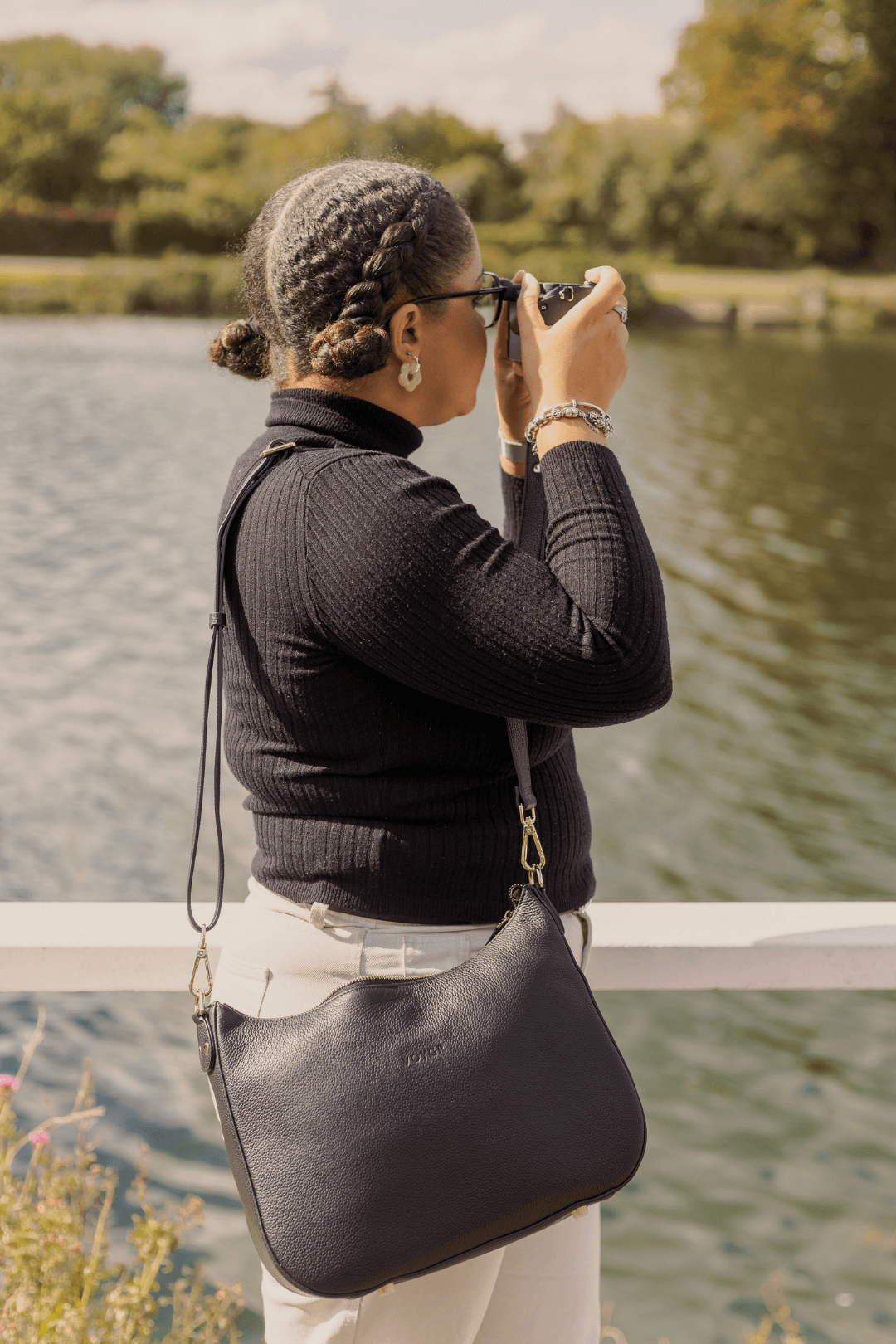 A woman with braided hair and glasses standing by a body of water, taking a photo with a camera. She is wearing a black turtleneck sweater, beige pants, and has a large black handbag hanging from her shoulder.