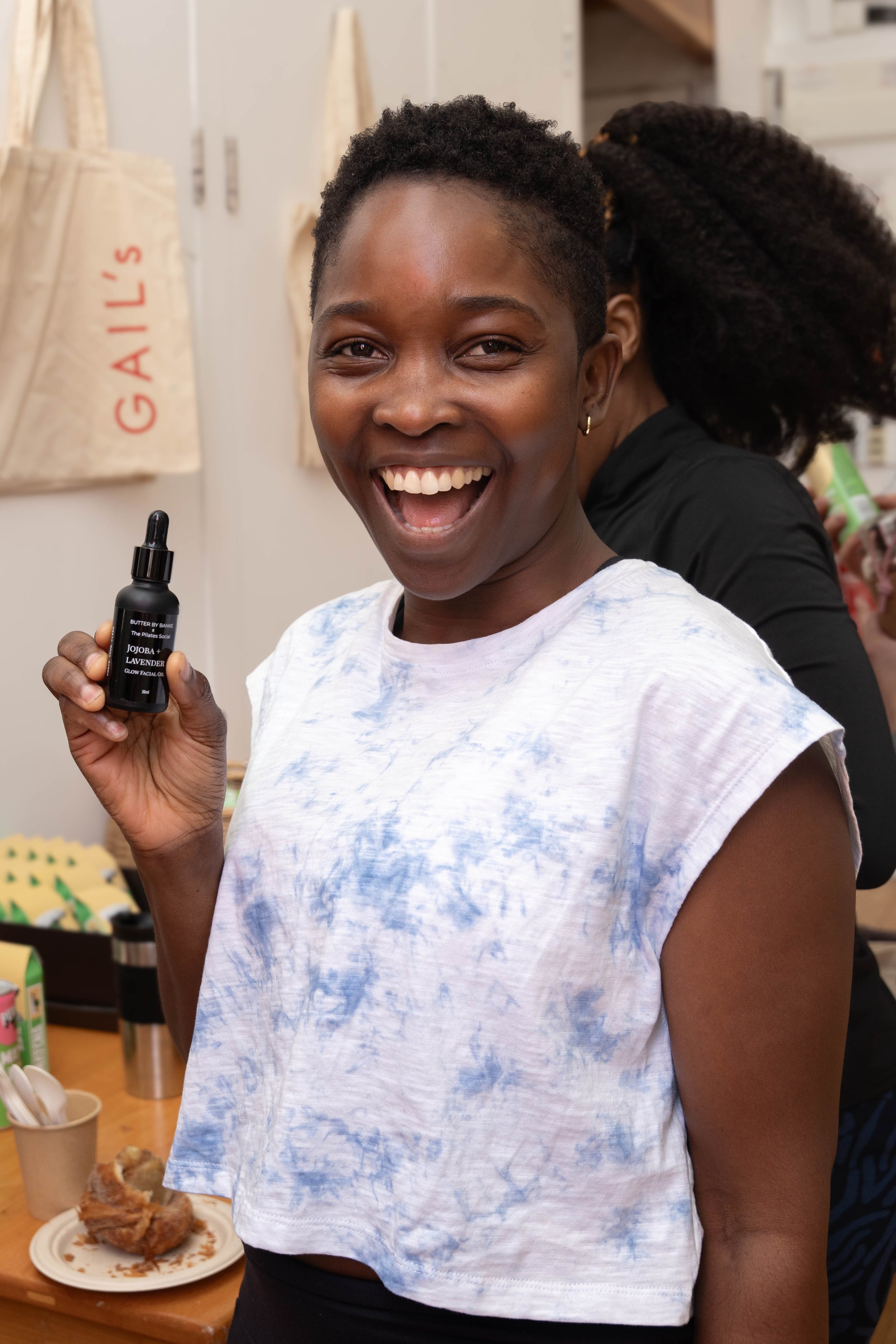 A smiling woman with short curly hair holding a dark dropper bottle labeled 'Jojoba + Lavender Glow Facial Oil'. She is wearing a white and blue tie-dye sleeveless shirt. In the background, there are beige tote bags hanging on the wall and a table wi