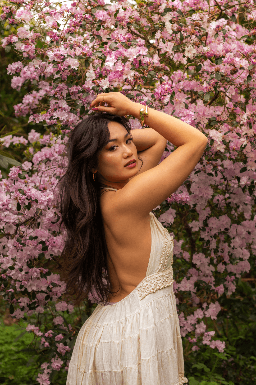 A woman with long, dark wavy hair standing in front of a flowering pink bush, wearing a cream-colored dress with lace details, jewelry, and looking confidently at the camera.