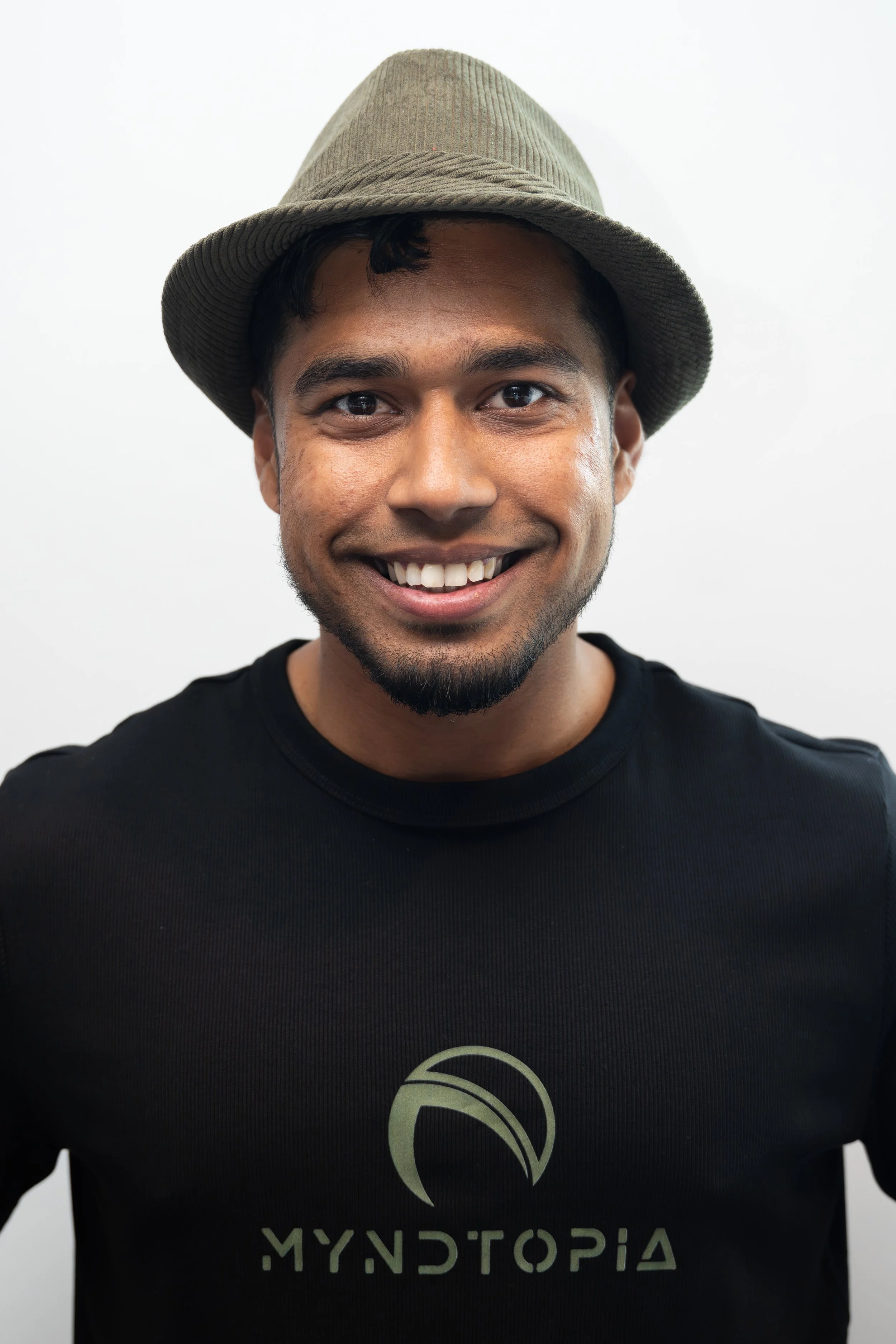 A young man with dark hair and a beard, wearing a light brown fedora hat and a black T-shirt with a logo and the word 'MyStopia' printed on it, smiling in front of a plain white background.
