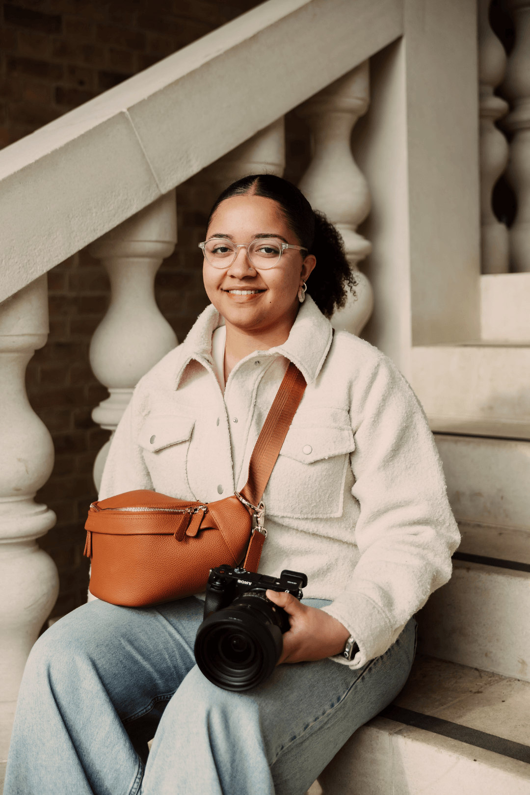 A woman sitting on stairs holding a camera, wearing glasses, a cream-colored jacket, and a brown crossbody bag.
