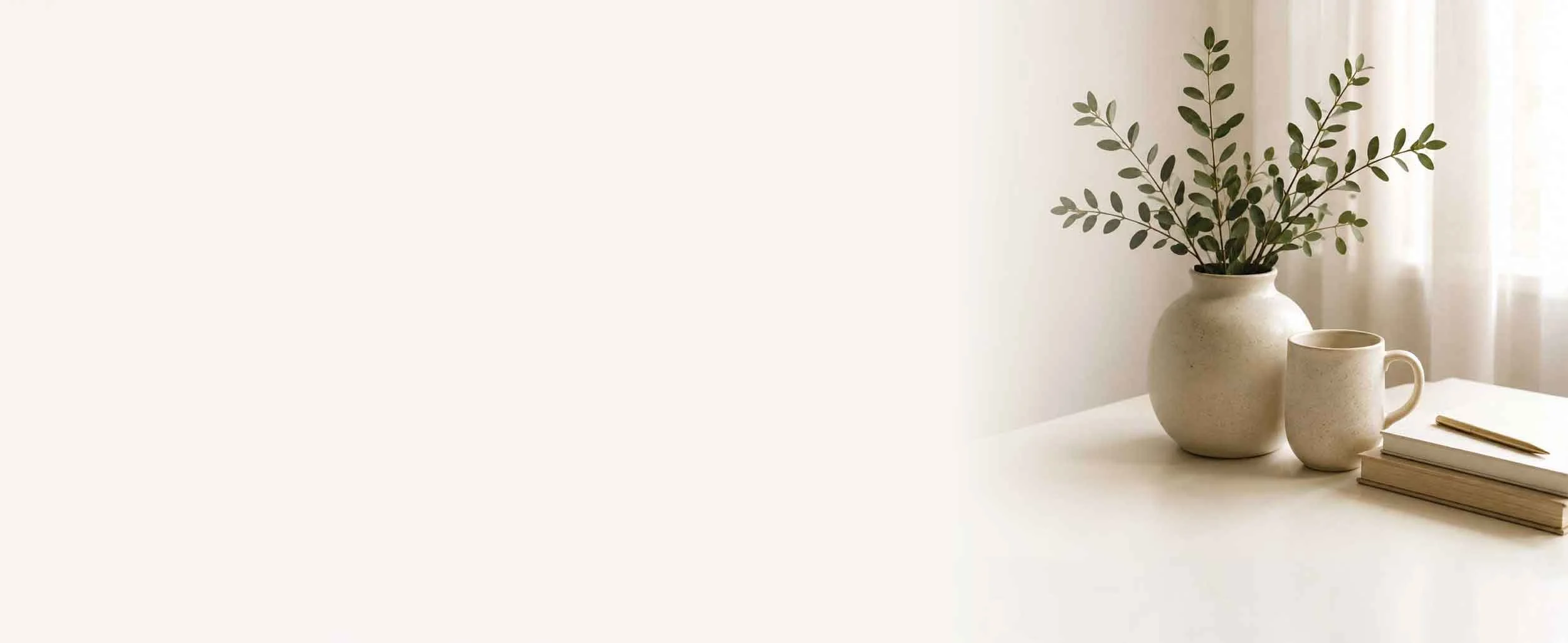 A minimalist arrangement of two beige ceramic vessels, one containing green leafy branches, placed on a white surface next to a closed handbook and a gold pen, with sheer curtains in the background.