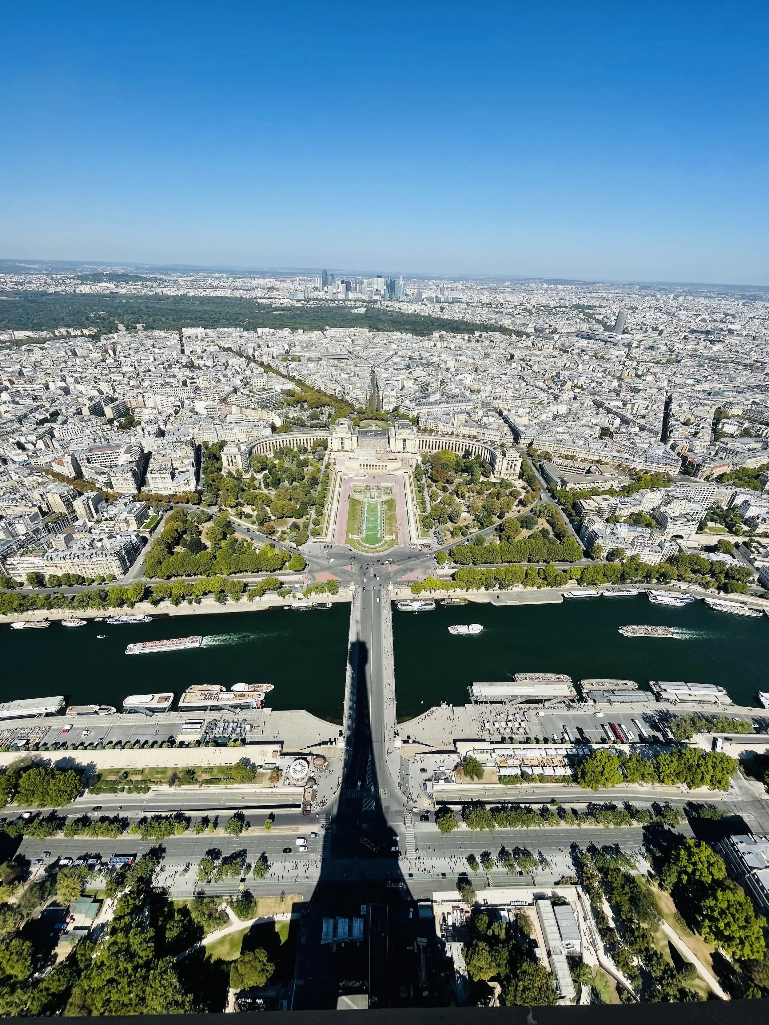 Vista aérea do Parque do Champ de Mars com a Torre Eiffel ao centro, com o rio Sena e barcos navegando, e a cidade de Paris ao fundo em dia claro.