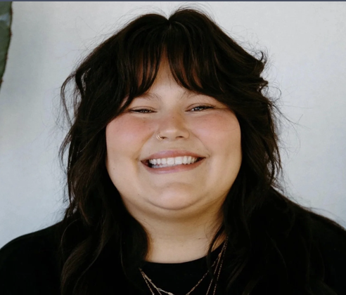 Smiling woman with shoulder-length dark hair and bangs, wearing a black top and layered necklaces, against a plain background.