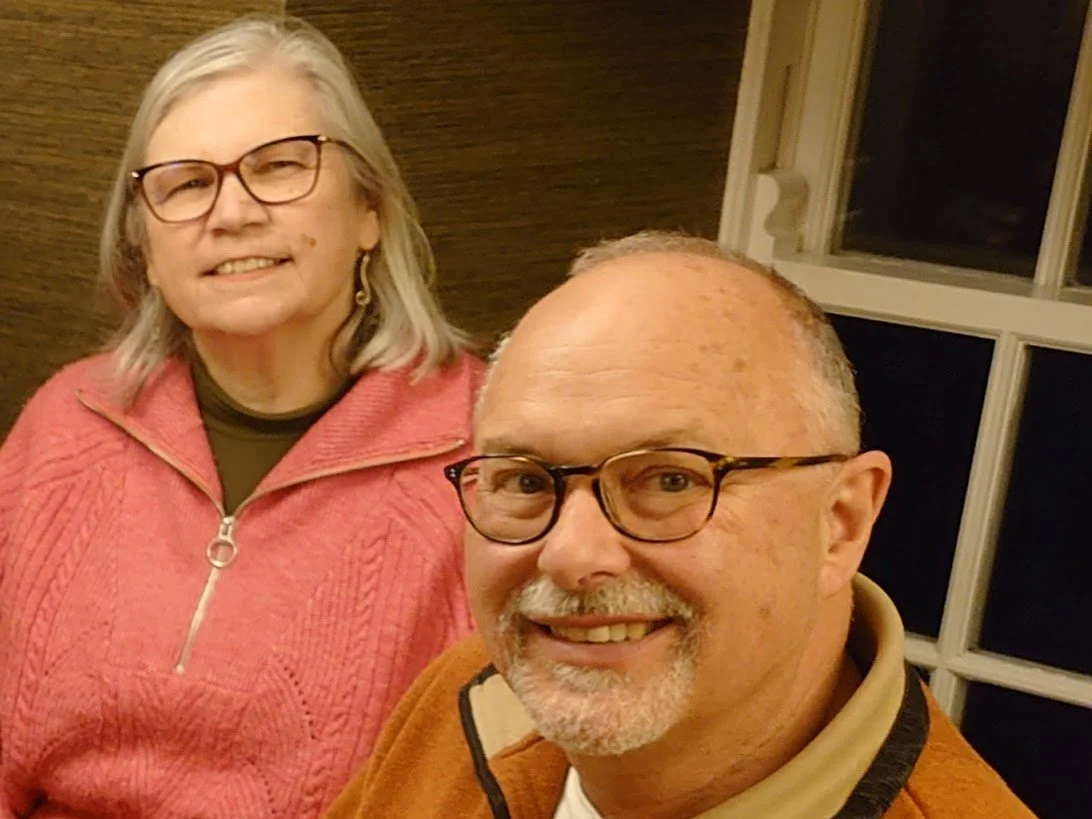 A smiling older man with glasses and a beard in the foreground, and an older woman with glasses and earrings in the background, both indoors at night.