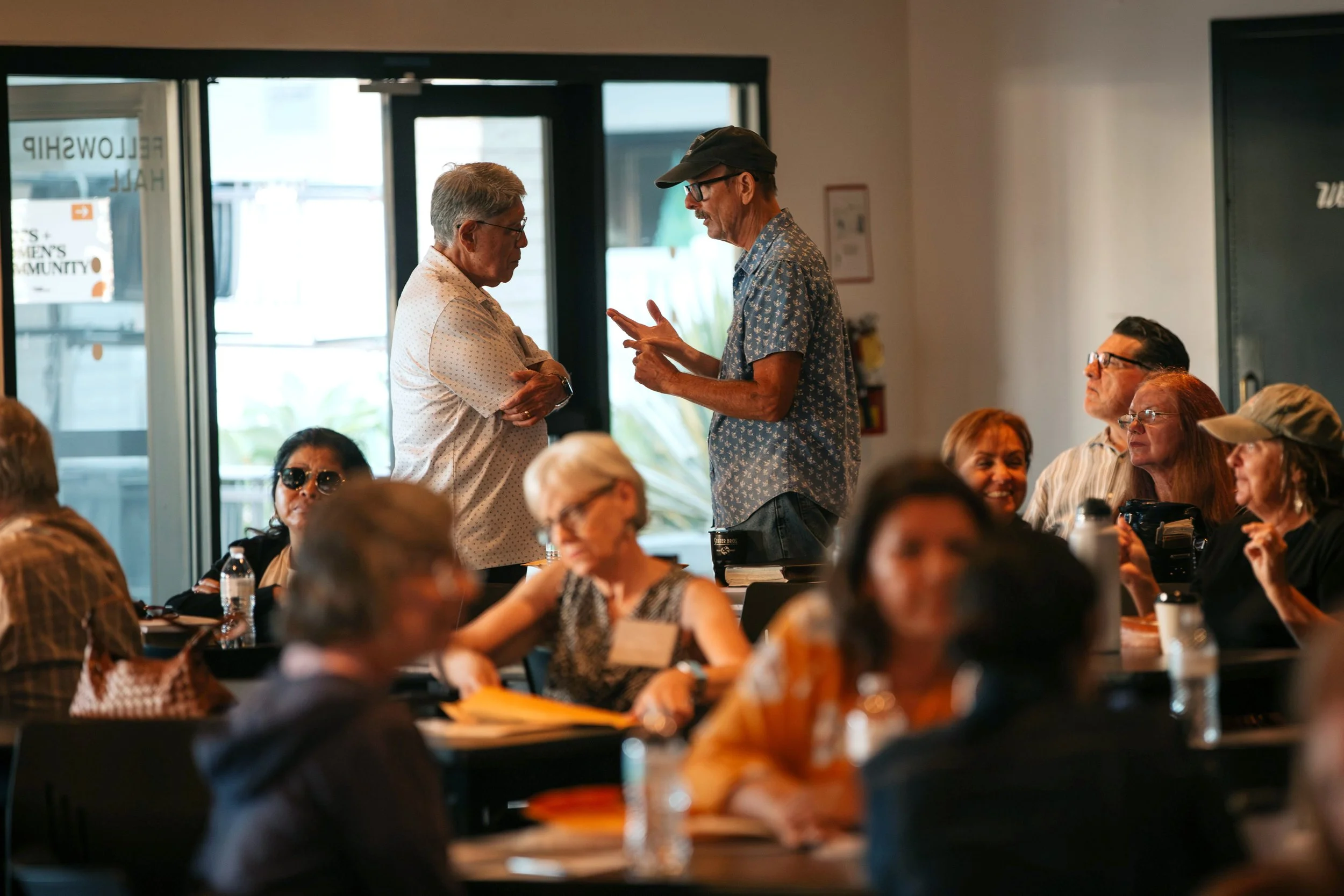 A man in a blue shirt and black cap shows something on his phone to an older woman in a white shirt during a gathering, with seated people around them in an indoor setting.