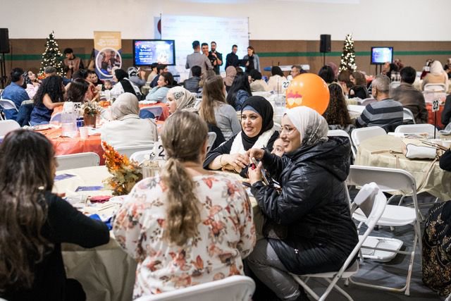 Women and men sitting at round tables in a large hall, attending an event with a panel of speakers on stage, decorated with Christmas trees and holiday decorations.